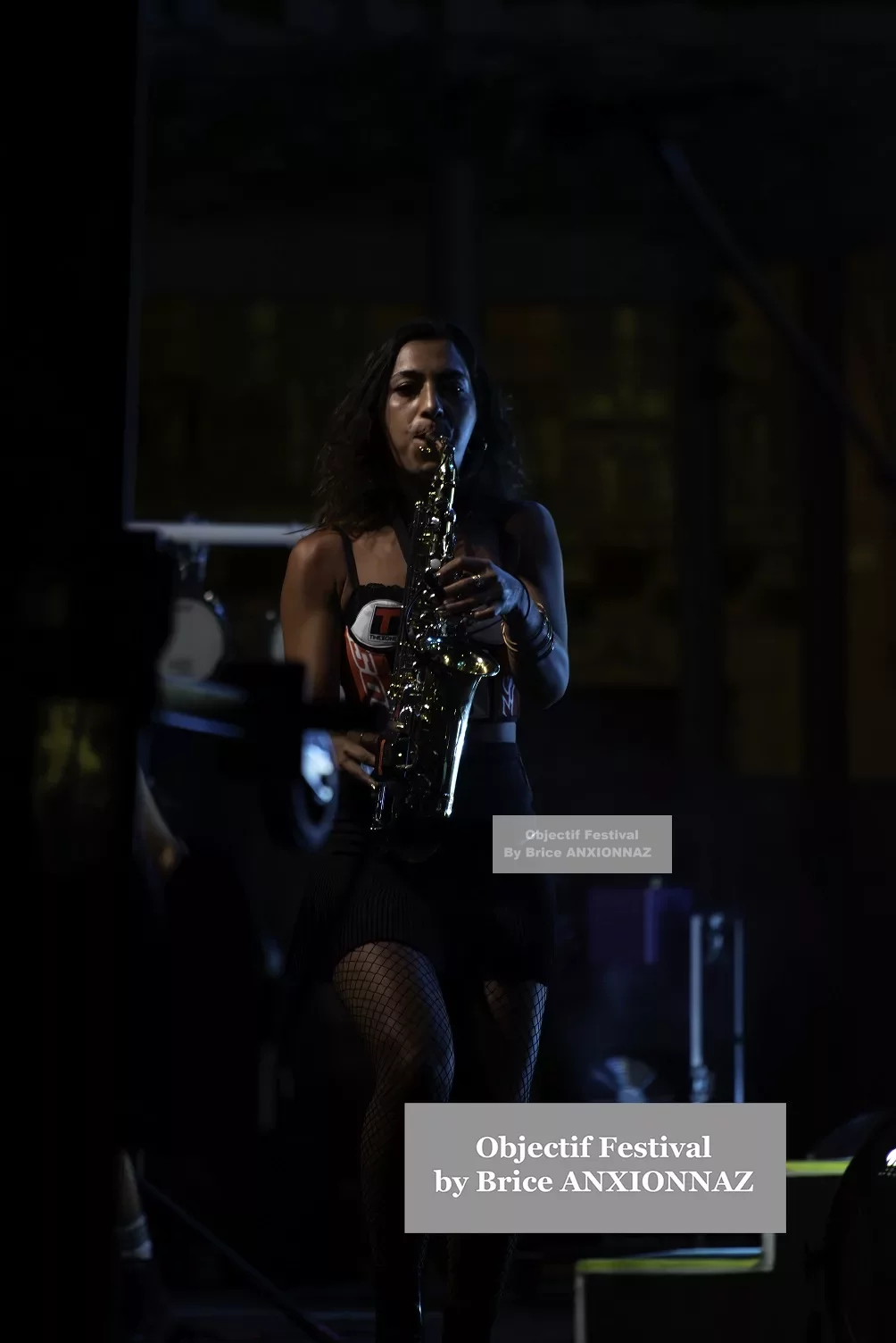 Une personne joue du saxophone sur scène, vêtue d'un haut noir et d'une jupe courte, dans une ambiance sombre avec des lumières tamisées et des ombres. Lej, Hyeres (06/08/2024)