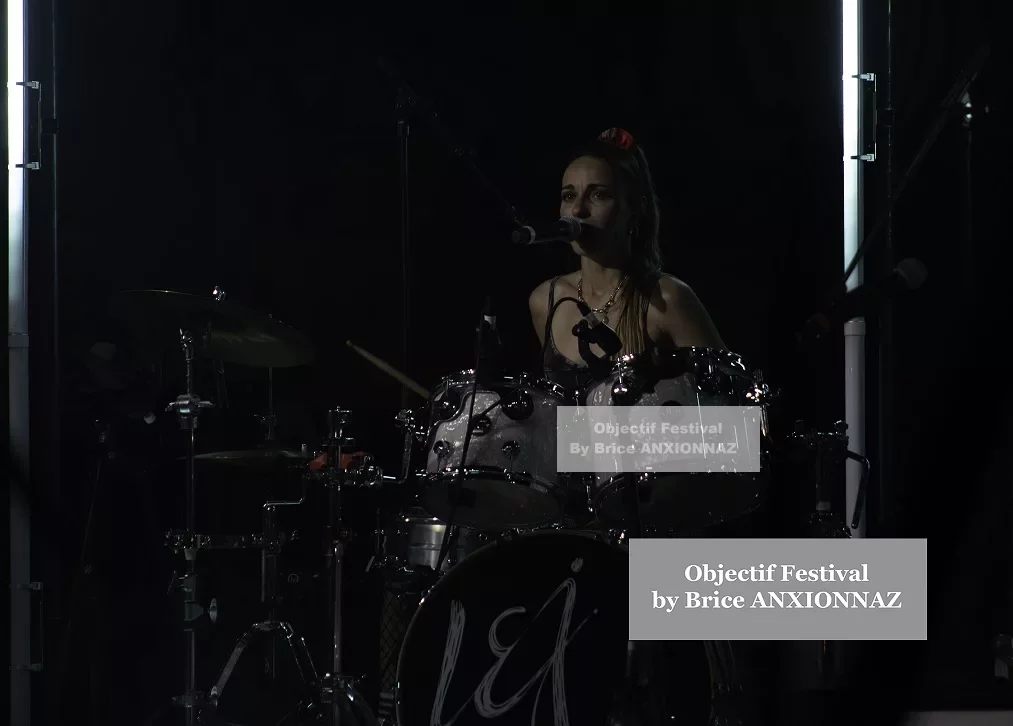 Une musicienne joue de la batterie sur scène, éclairée par des lumières vives, dans une ambiance sombre et dynamique, avec des instruments en métal brillant et un fond noir. Lej, Hyeres (06/08/2024)