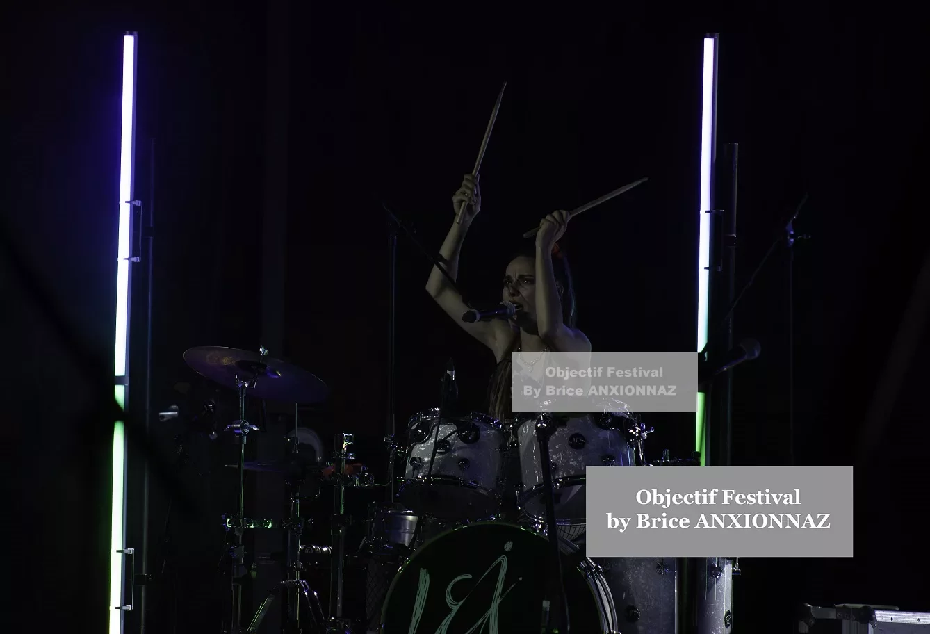 Une personne joue de la batterie sur scène, entourée de lumières colorées, dans une ambiance sombre et dynamique, avec des éléments de percussion brillants en arrière-plan. Lej, Hyeres (06/08/2024)