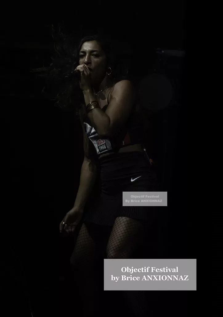 Une artiste chante sur scène, vêtue d'une tenue noire et colorée, avec des lumières tamisées créant une ambiance dynamique et énergique. Lej, Hyeres (06/08/2024)