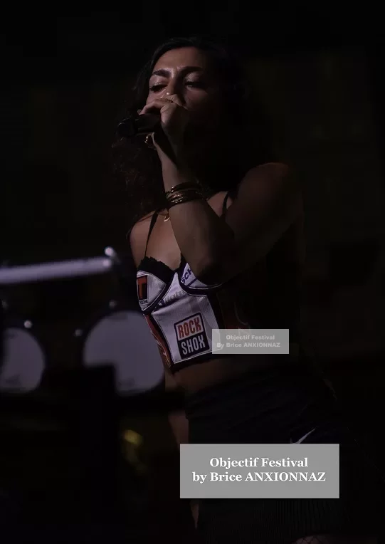 Une artiste chante sur scène, vêtue d'un haut coloré et d'une jupe noire, avec un éclairage tamisé créant une ambiance dynamique et énergique. Lej, Hyeres (06/08/2024)