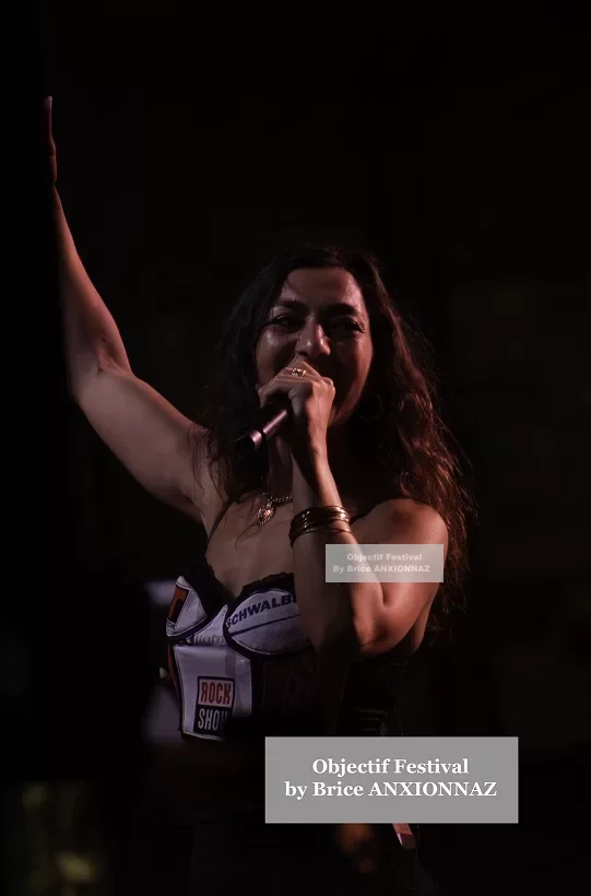 Une personne chante sur scène, vêtue d'une tenue noire et blanche, avec des lumières tamisées créant une ambiance dynamique et énergique. Lej, Hyeres (06/08/2024)