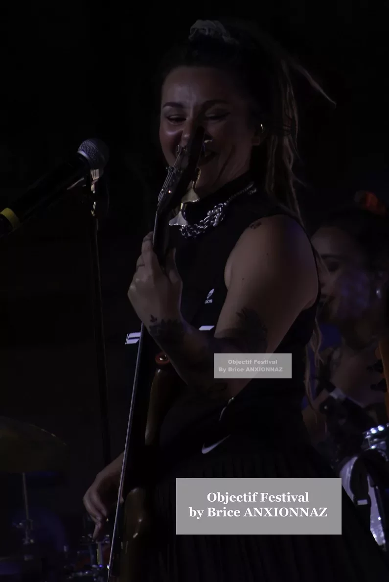 Une musicienne souriante joue de la basse sur scène, vêtue d'une tenue noire, avec des éclairages tamisés créant une ambiance dynamique et festive. Lej, Hyeres (06/08/2024)