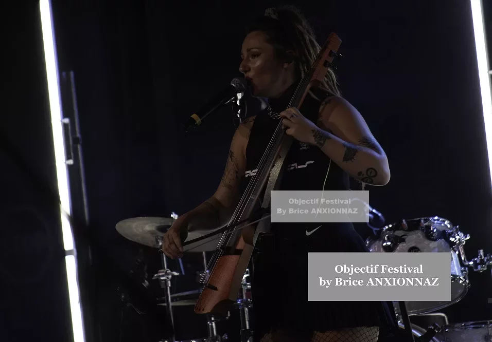 Une personne chante sur scène, tenant un instrument de musique, avec des éclairages modernes en arrière-plan et une ambiance dynamique, vêtue d'une tenue noire. Lej, Hyeres (06/08/2024)