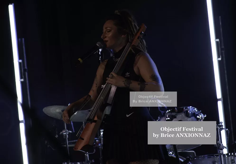 Une personne chante sur scène, tenant un instrument, avec des lumières vives en arrière-plan et une ambiance dynamique, vêtue d'une tenue noire et sportive. Lej, Hyeres (06/08/2024)