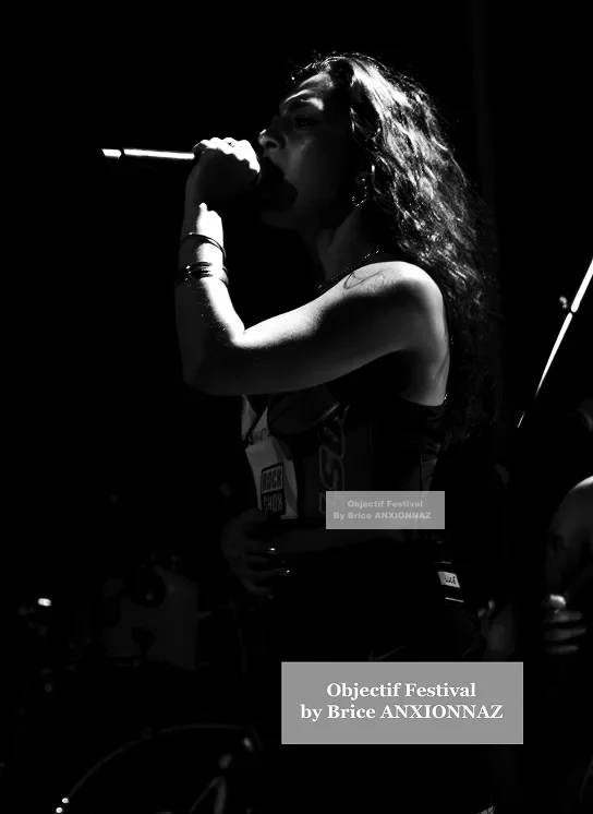 Une personne chante sur scène, éclairée par un éclairage dramatique, créant un contraste fort avec l'arrière-plan sombre, tandis que des instruments de musique sont visibles en arrière-plan. Lej, Hyeres (06/08/2024)