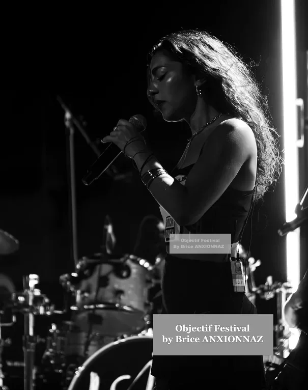 Une artiste chante sur scène, éclairée par des lumières vives, avec un fond de batterie, dans une ambiance monochrome et dynamique. Lej, Hyeres (06/08/2024)