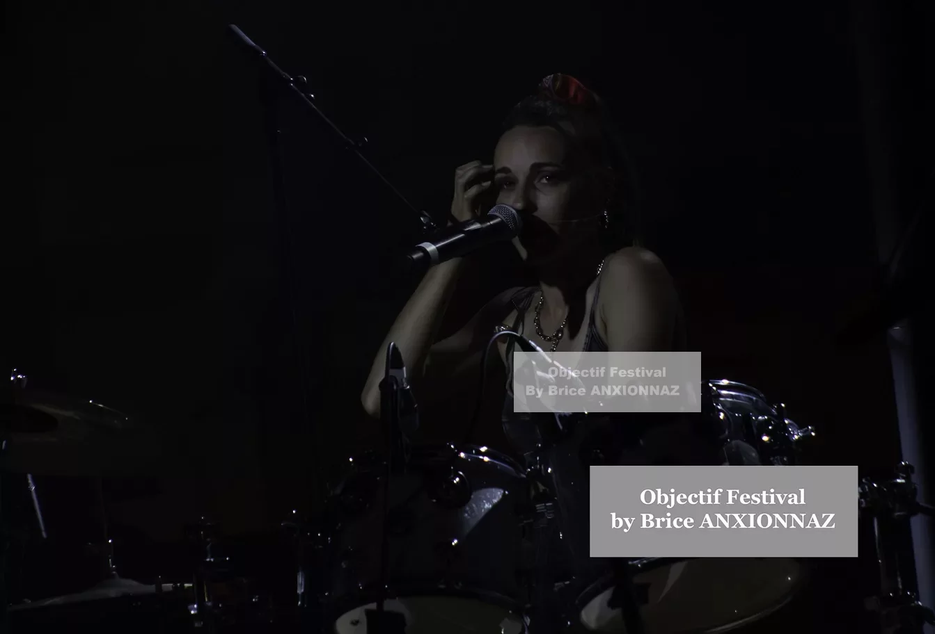 Une musicienne est assise derrière une batterie, tenant un micro, dans une ambiance sombre avec des éclairages tamisés, créant une atmosphère intimiste au festival. Lej, Hyeres (06/08/2024)
