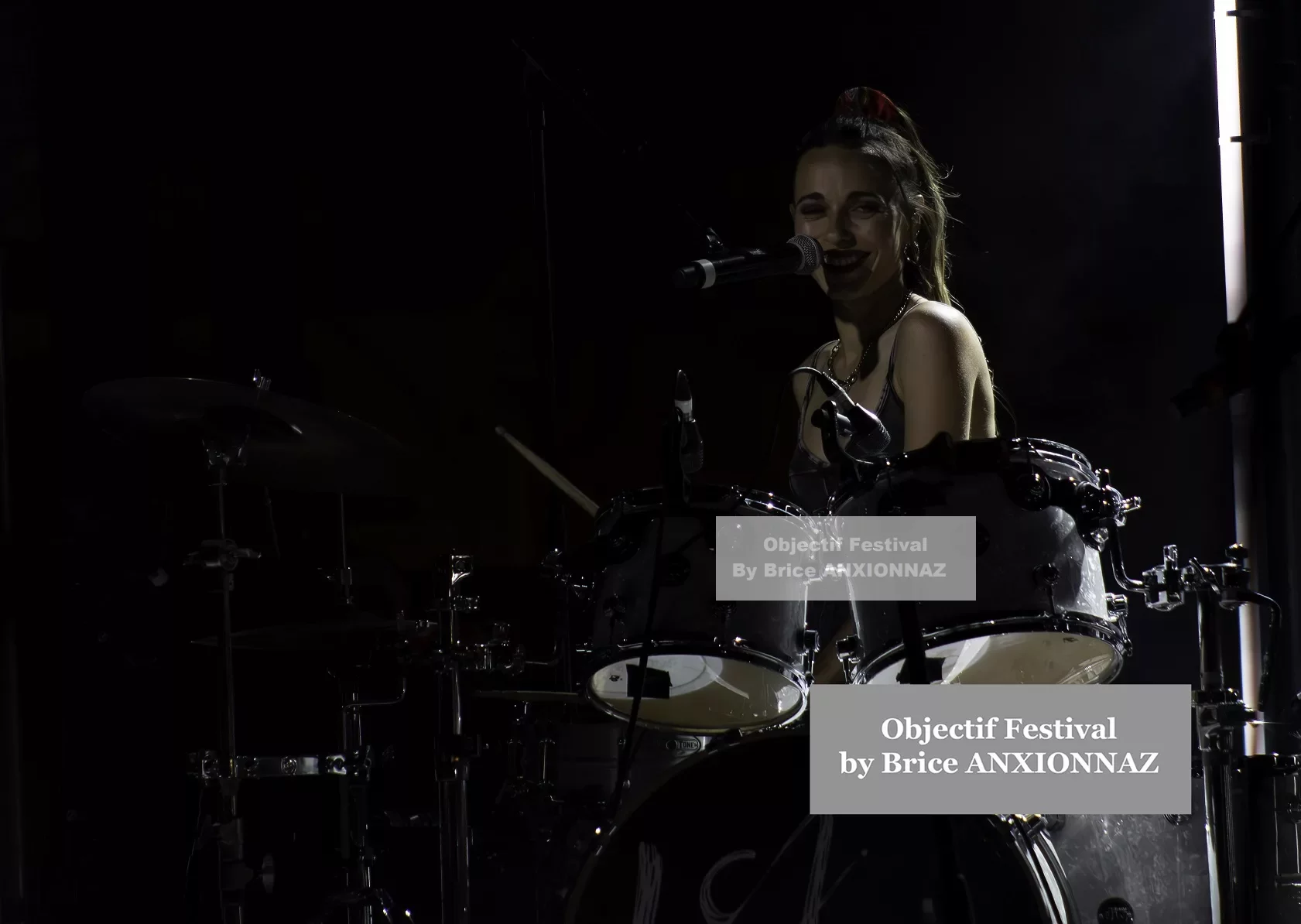 Une musicienne souriante joue de la batterie sur scène, éclairée par des lumières tamisées, créant une ambiance dynamique et festive. Lej, Hyeres (06/08/2024)