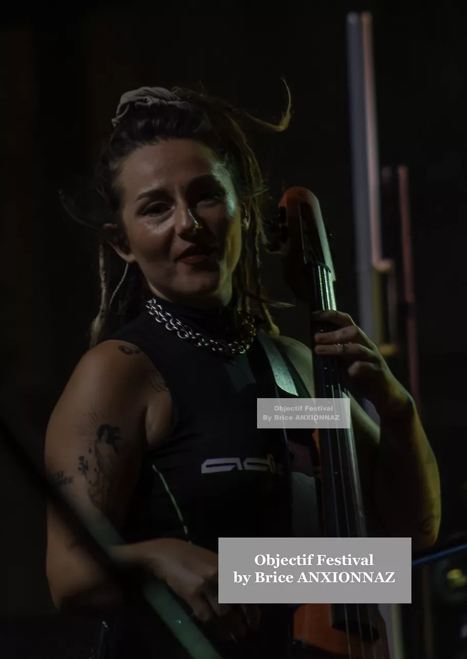 Une musicienne joue du violoncelle sur scène, éclairée par des lumières tamisées, portant un haut noir et des accessoires argentés, avec une ambiance dynamique et festive. Lej, Hyeres (06/08/2024)