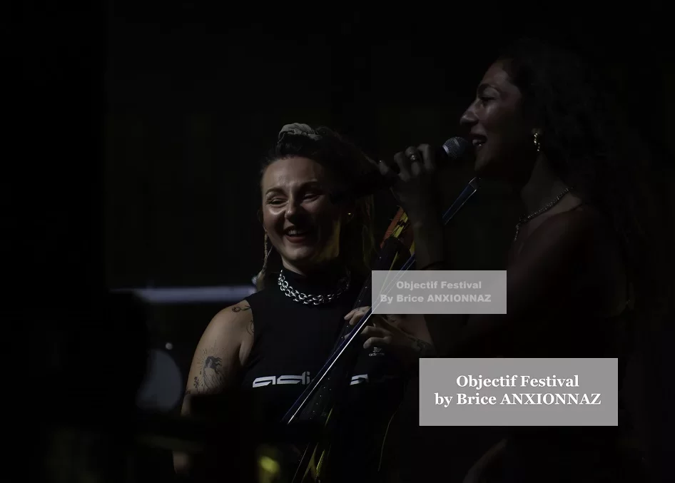 Deux chanteuses souriantes se produisent sur scène, éclairées par des lumières tamisées, avec des vêtements noirs et des accessoires brillants, créant une ambiance festive et dynamique. Lej, Hyeres (06/08/2024)