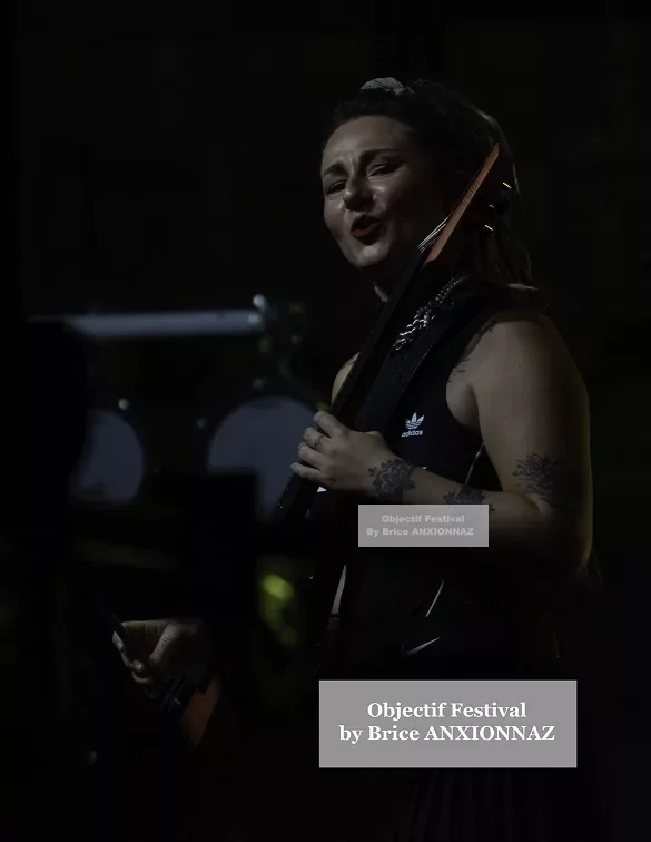 Une musicienne souriante joue de la guitare sur scène, éclairée par des lumières tamisées, avec des instruments flous en arrière-plan, créant une ambiance dynamique et festive. Lej, Hyeres (06/08/2024)