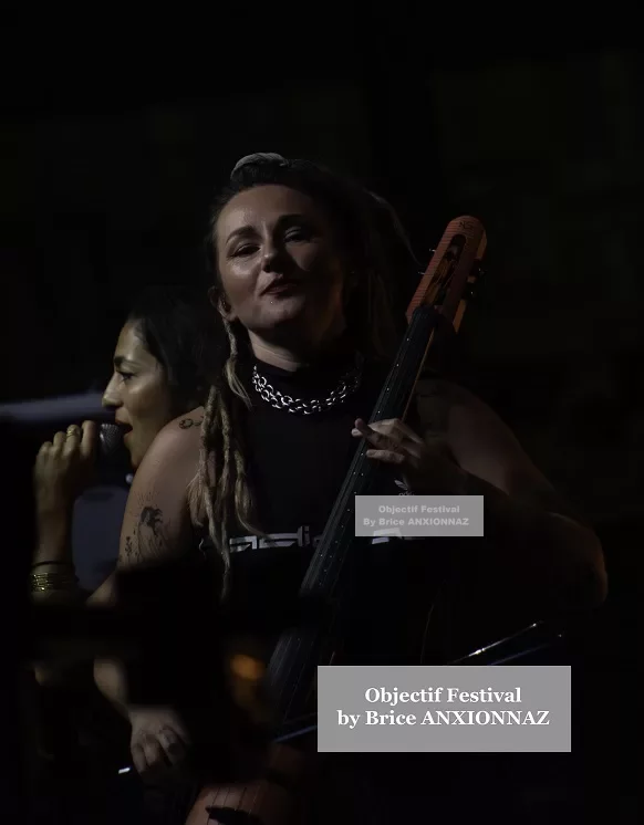 Une musicienne souriante joue d'un instrument à cordes sur scène, tandis qu'une autre chante en arrière-plan, sous un éclairage tamisé qui crée une ambiance intime. Lej, Hyeres (06/08/2024)