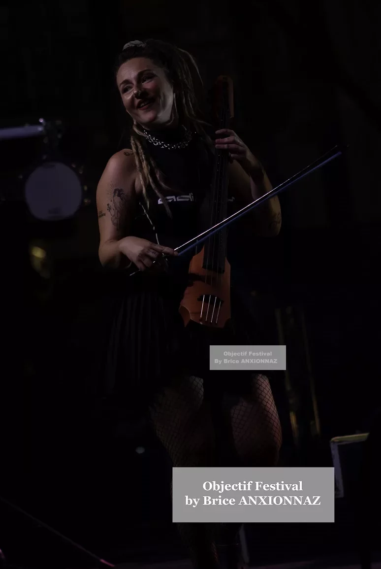 Une personne souriante joue d'un instrument à cordes sur scène, vêtue d'une tenue noire et de collants résille, sous un éclairage tamisé qui crée une ambiance festive. Lej, Hyeres (06/08/2024)