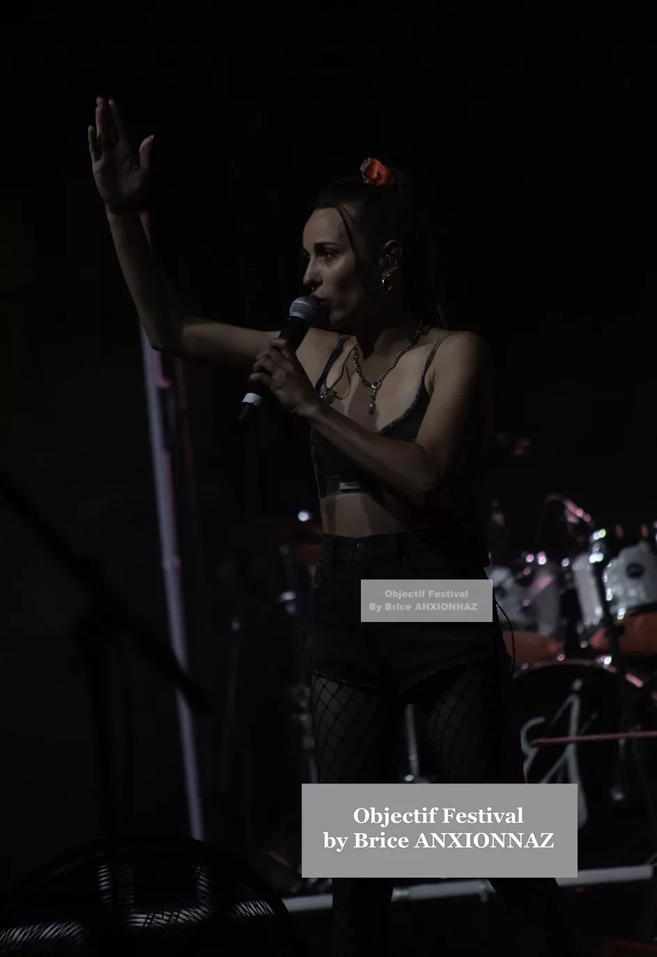 Une artiste sur scène, vêtue d'un haut noir et de shorts, chante dans un éclairage tamisé, levant une main pour interagir avec le public, tandis qu'un groupe de musique est visible en arrière-plan. Lej, Hyeres (06/08/2024)