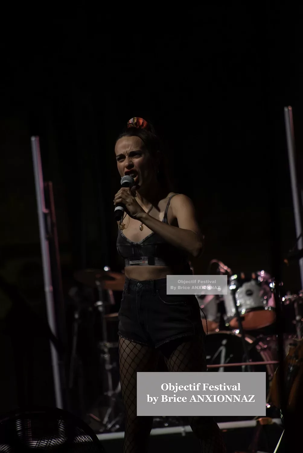 Une personne chante sur scène, vêtue d'un haut noir et de shorts, avec des collants en résille, devant un ensemble de batterie, dans une ambiance sombre et lumineuse. Lej, Hyeres (06/08/2024)
