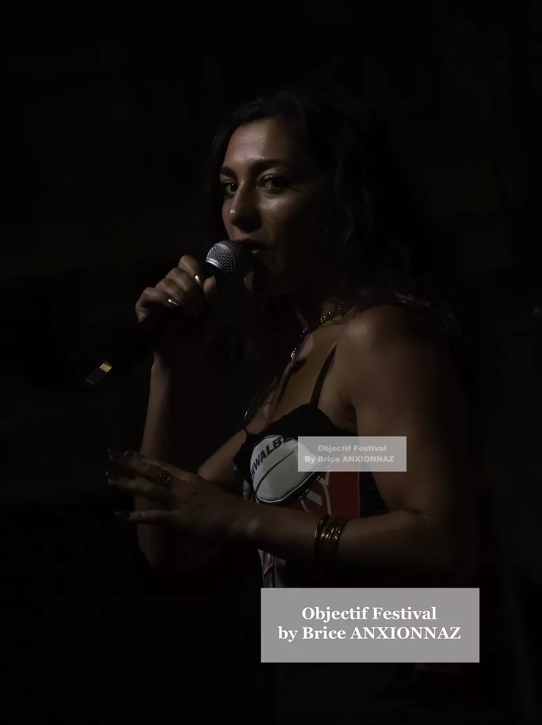 Une personne chante dans un éclairage tamisé, portant un haut coloré, avec un microphone à la main, créant une ambiance dynamique et captivante. Lej, Hyeres (06/08/2024)