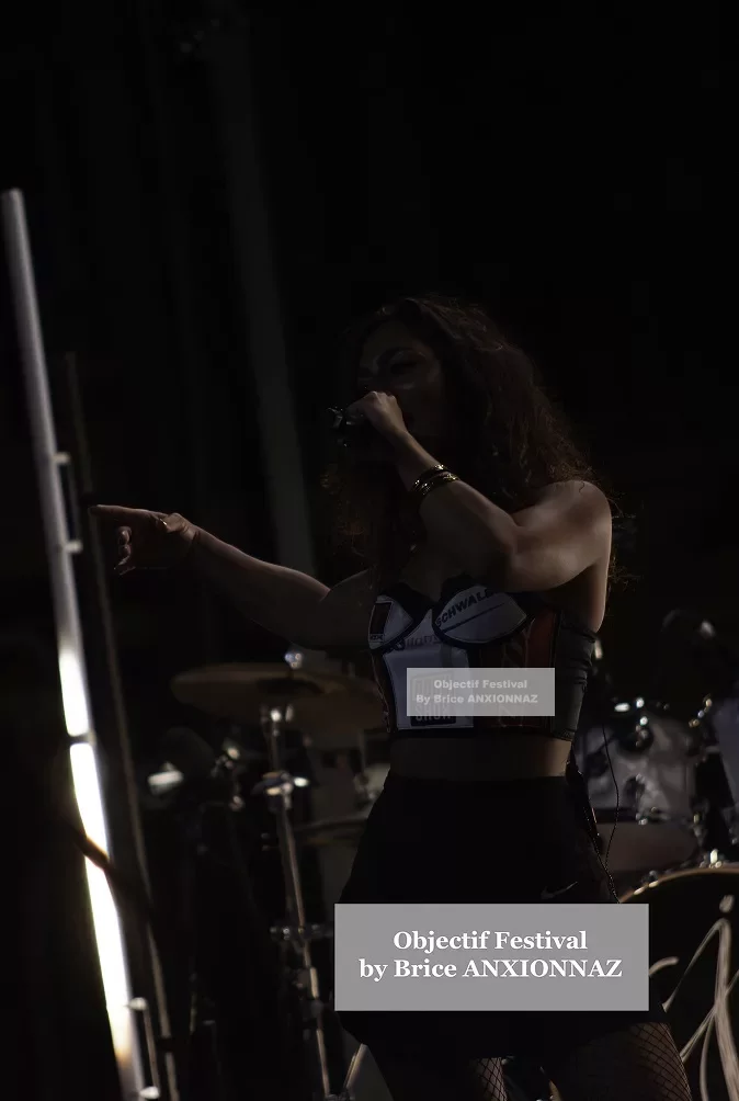 Une artiste chante sur scène, vêtue d'un haut noir et d'une jupe, avec des lumières tamisées en arrière-plan, créant une ambiance dynamique et énergique. Lej, Hyeres (06/08/2024)