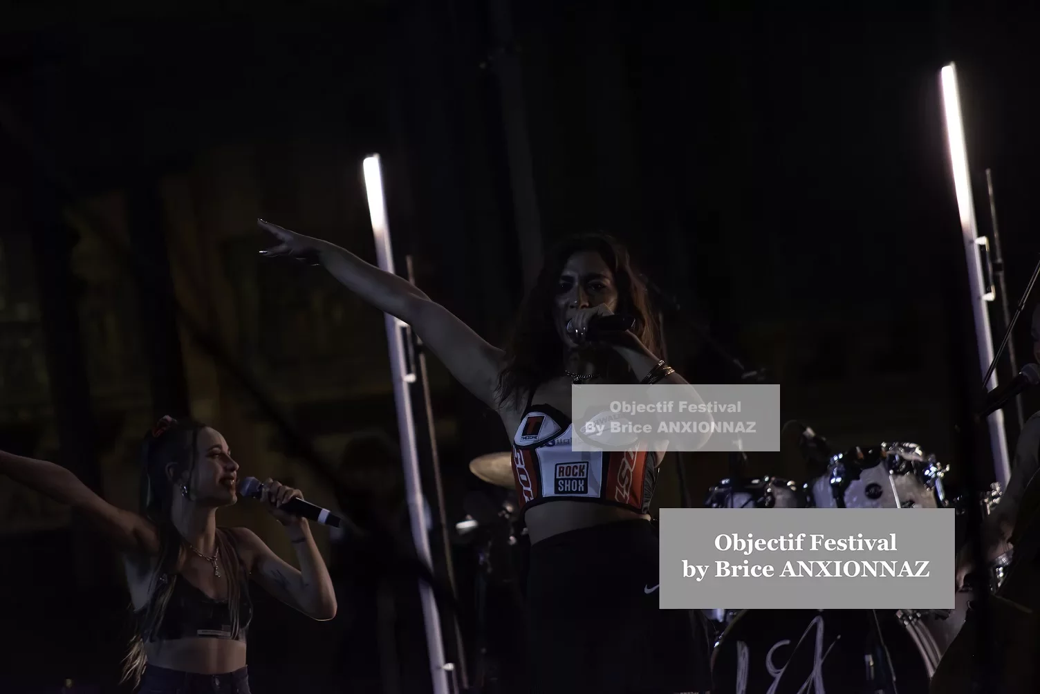 Deux artistes se produisent sur scène, l'une chantant avec un micro, l'autre levant le bras. L'éclairage est tamisé, créant une ambiance dynamique et énergique. Lej, Hyeres (06/08/2024)