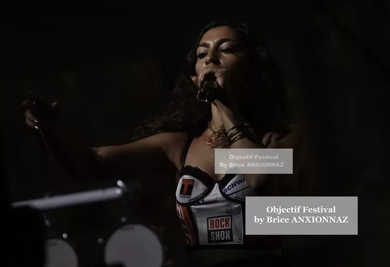 Une personne chante sur scène, vêtue d'un haut coloré, avec des lumières tamisées en arrière-plan, créant une ambiance dynamique et énergique. Lej, Hyeres (06/08/2024)