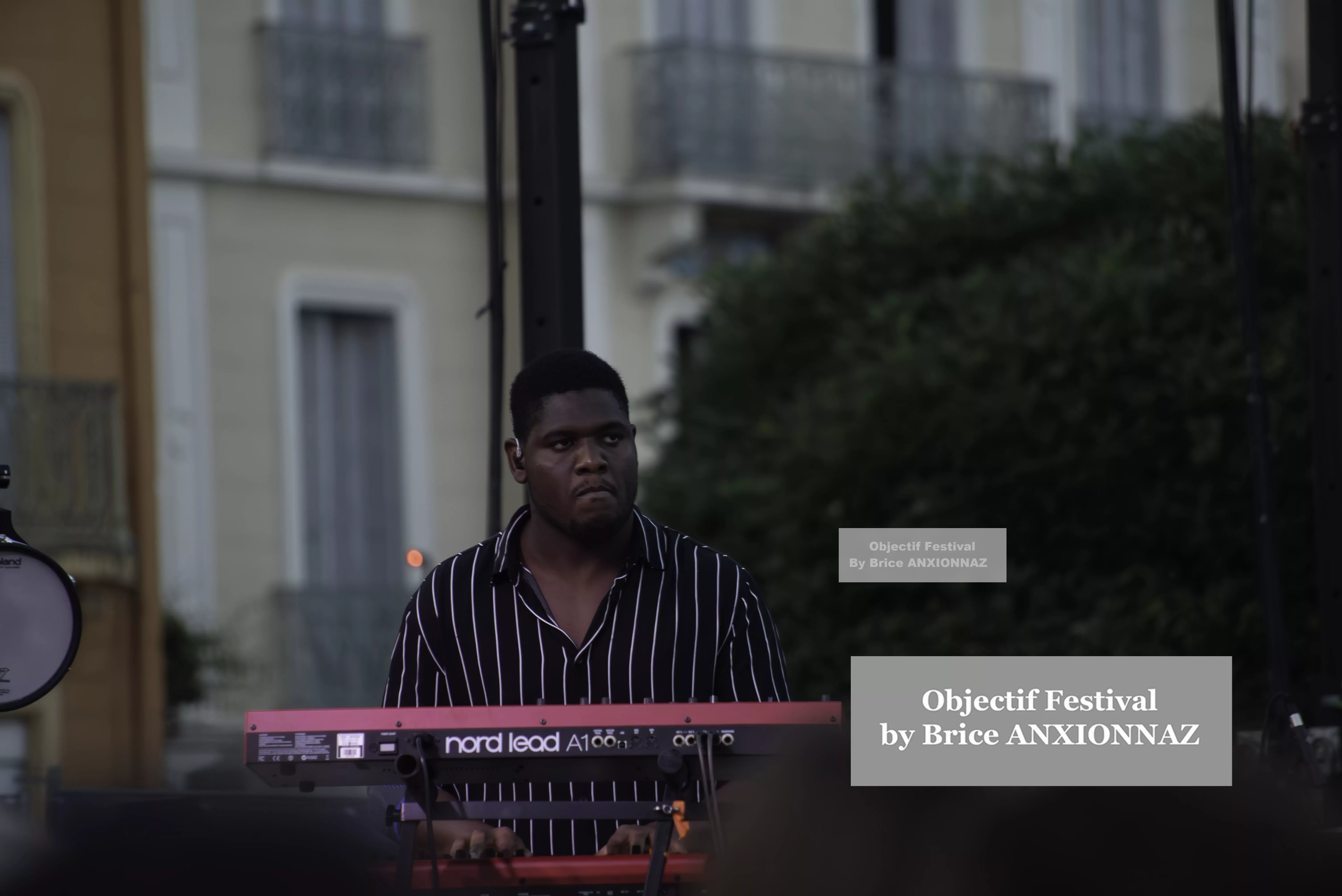 Un musicien joue du clavier sur scène, vêtu d'une chemise à rayures, avec un fond de bâtiments et de verdure, sous une lumière douce du soir. Itsouidad, Hyeres (06/08/2024)