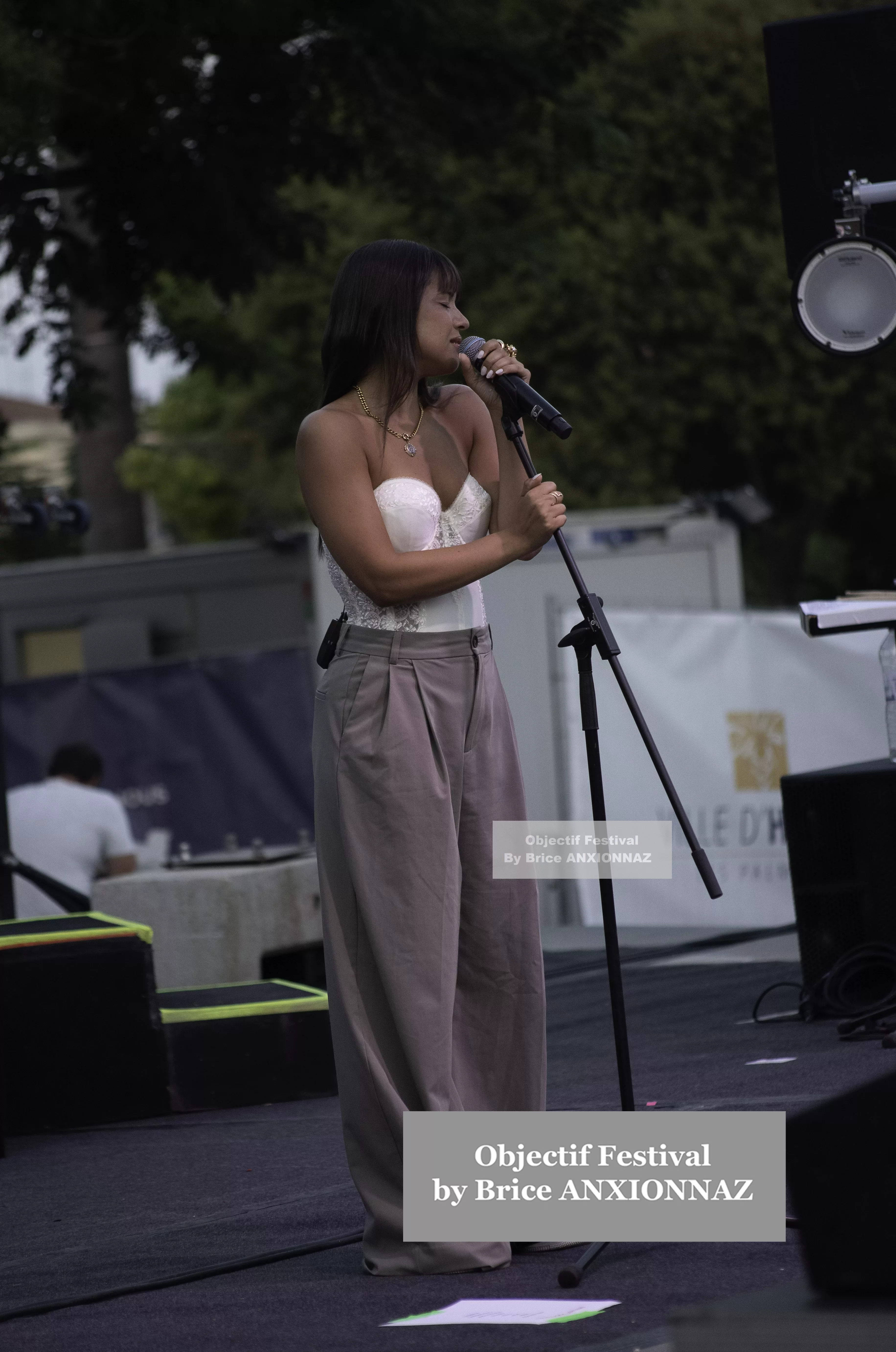 Une personne chante sur scène, vêtue d'un haut blanc et de pantalons beiges, sous un éclairage naturel, avec des arbres en arrière-plan et une ambiance festive. Itsouidad, Hyeres (06/08/2024)