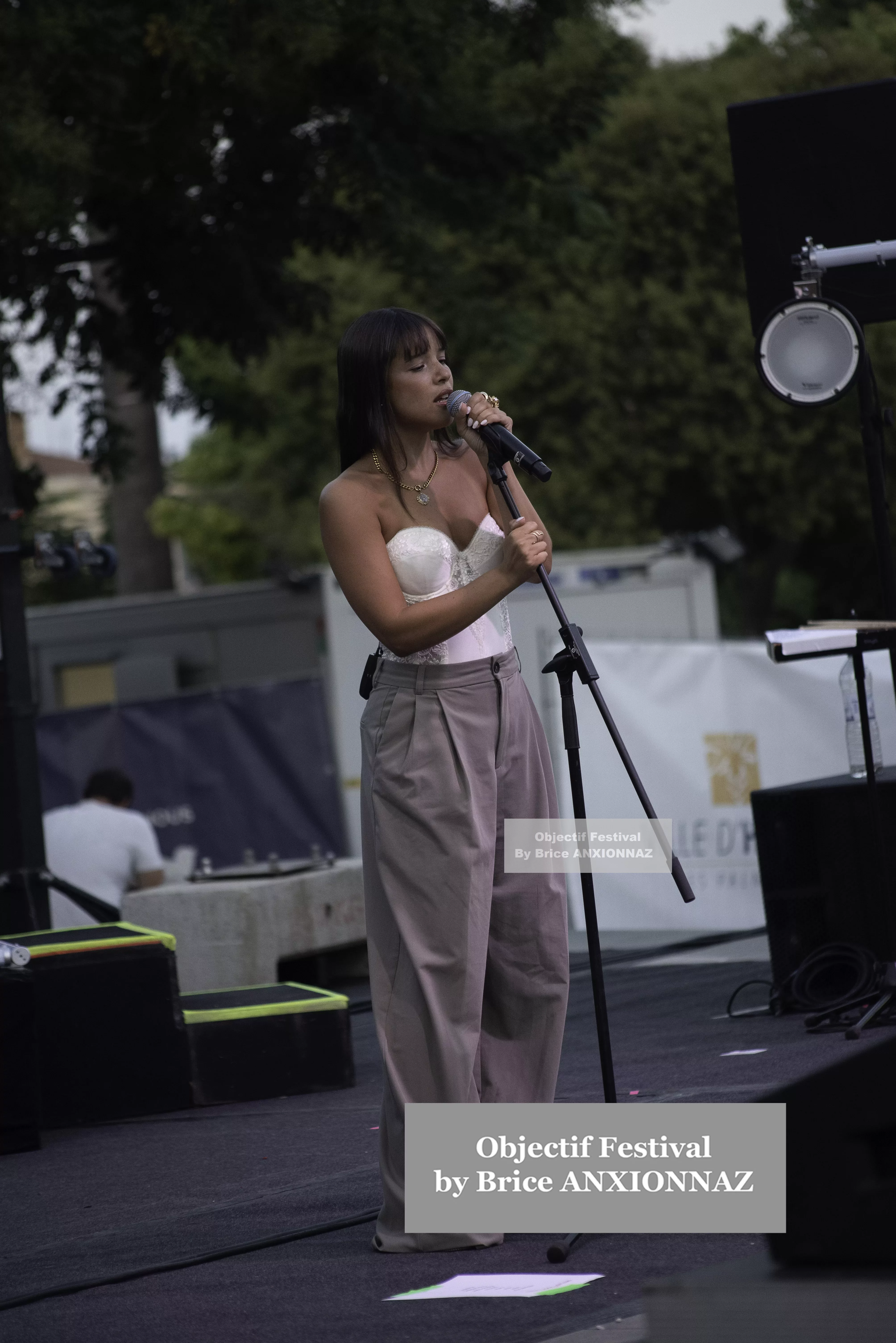 Une personne chante sur scène, vêtue d'un haut blanc et de pantalons larges, avec un microphone à la main, tandis qu'un public flou est visible en arrière-plan. Itsouidad, Hyeres (06/08/2024)