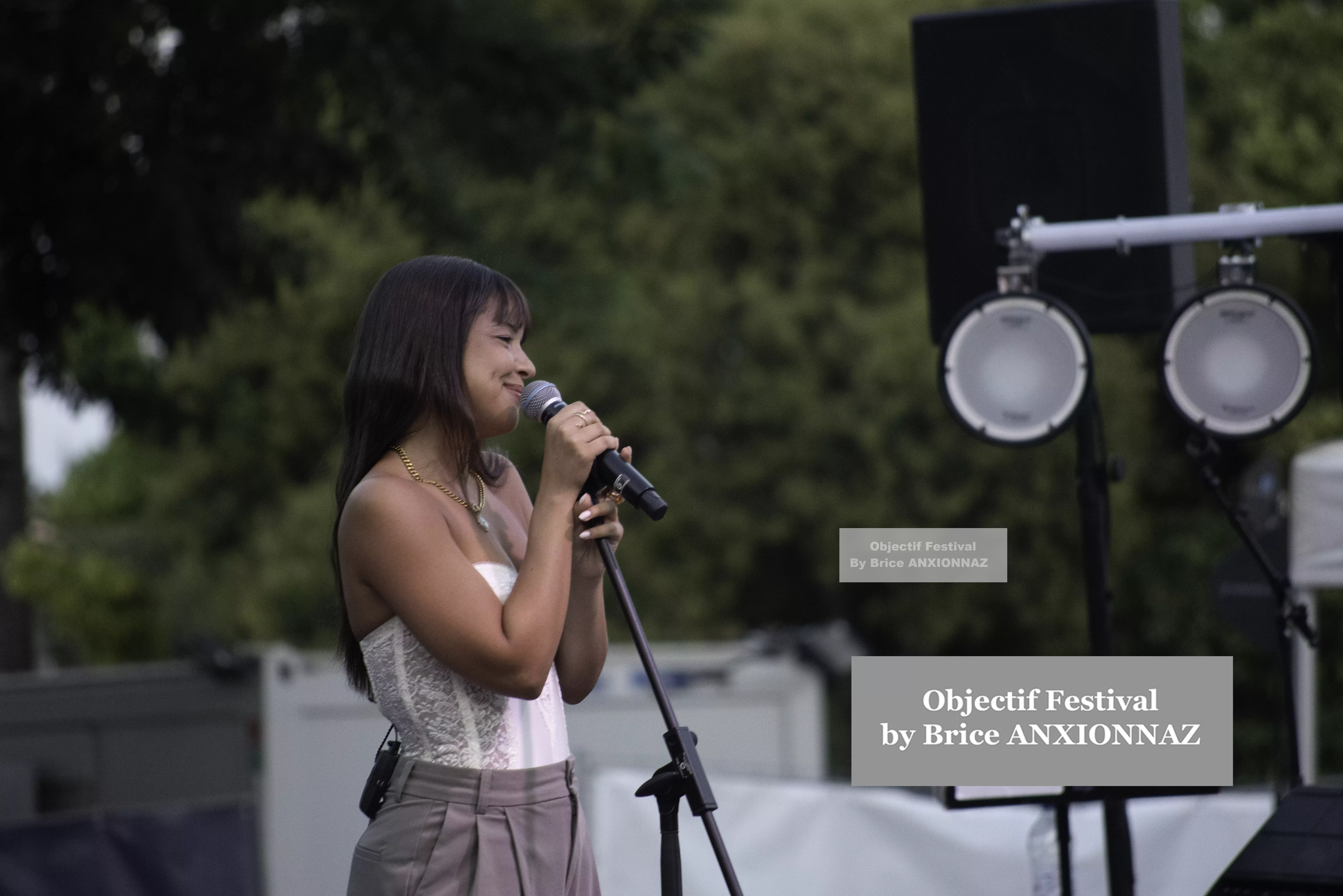 Une personne chante sur scène, vêtue d'un haut clair et d'un pantalon, avec des haut-parleurs en arrière-plan et un éclairage naturel, créant une ambiance festive. Itsouidad, Hyeres (06/08/2024)