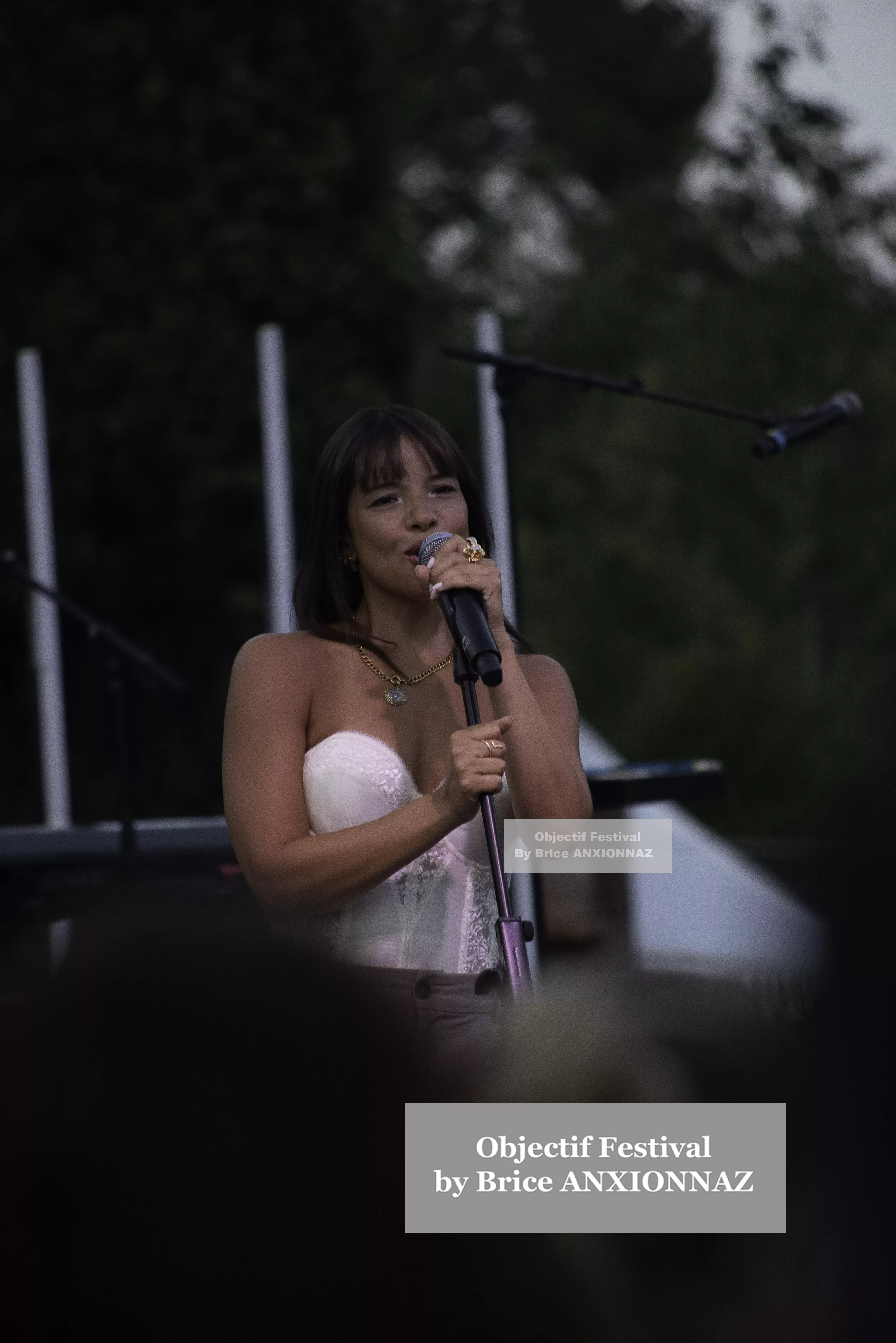 Une artiste chante sur scène, vêtue d'un haut blanc, avec un microphone à la main, tandis qu'un public flou l'écoute, sous une lumière douce et naturelle. Itsouidad, Hyeres (06/08/2024)
