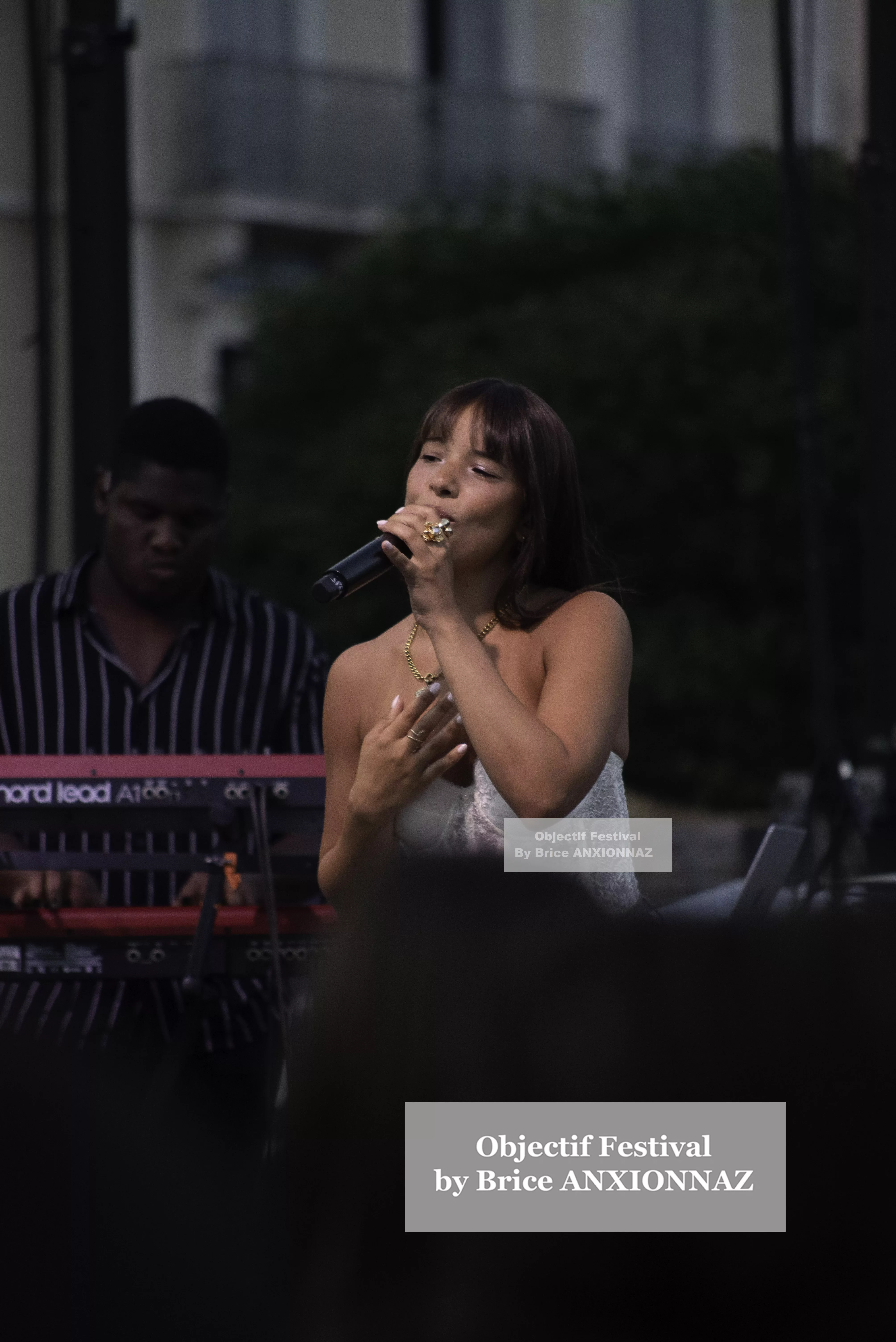 Une chanteuse en robe blanche se produit sur scène, tenant un micro, tandis qu'un musicien en arrière-plan joue des claviers, sous une lumière tamisée et une ambiance festive. Itsouidad, Hyeres (06/08/2024)