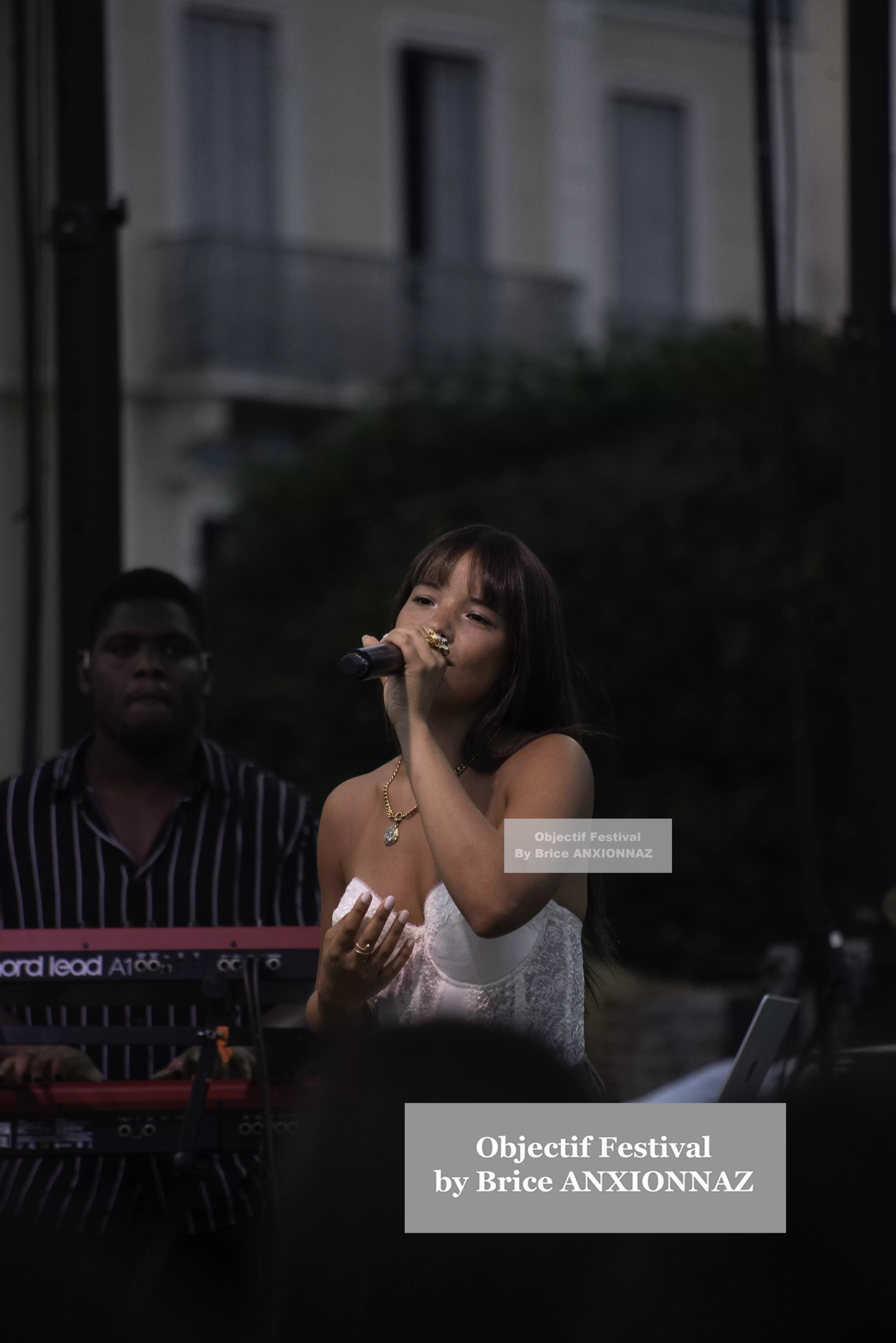 Une chanteuse en tenue blanche se produit sur scène, tenant un micro, tandis qu'un musicien en arrière-plan joue d'un clavier, sous une lumière tamisée. Itsouidad, Hyeres (06/08/2024)