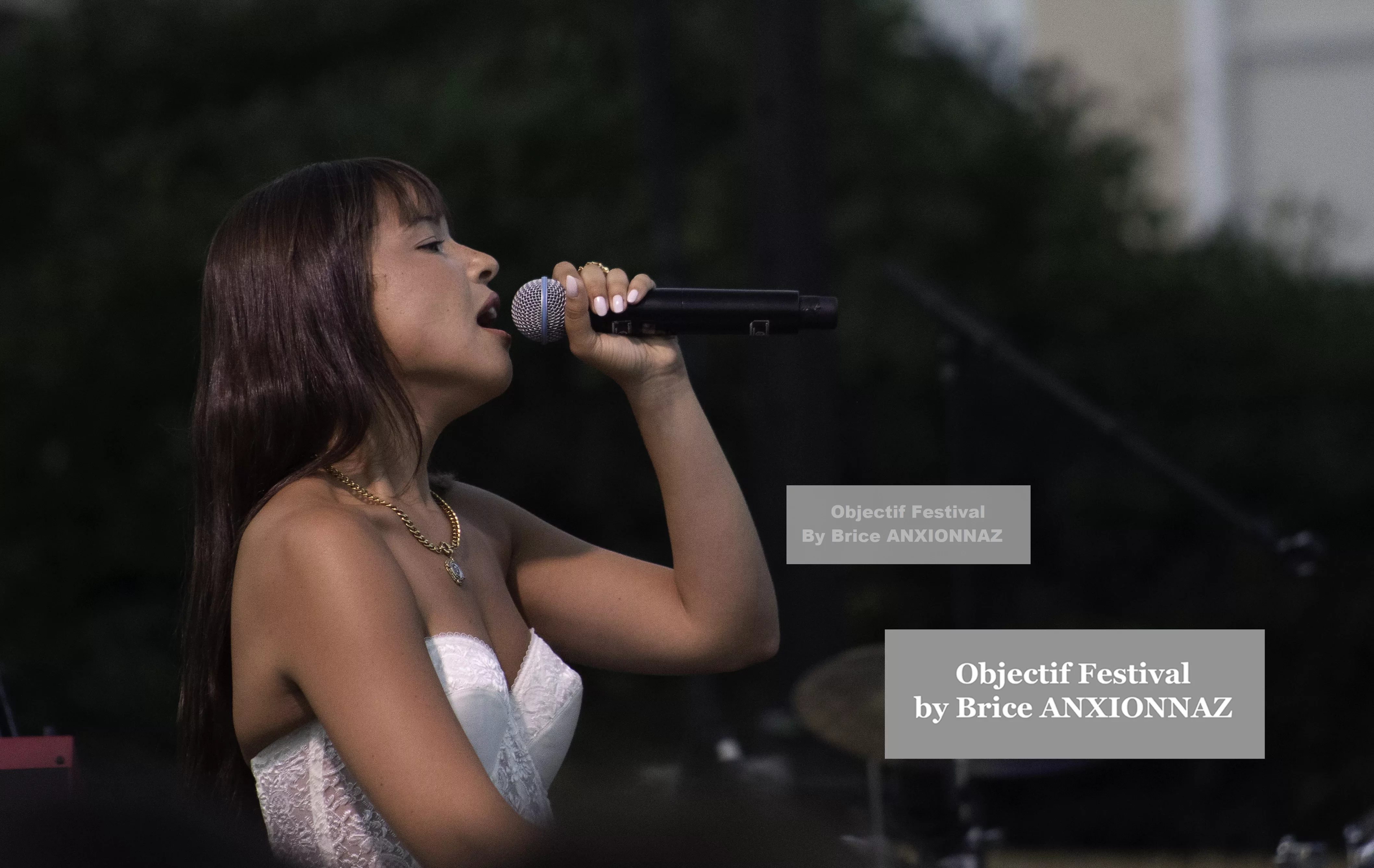 Une chanteuse interprète une chanson sur scène, vêtue d'une robe blanche, avec un microphone à la main, sous une lumière douce, entourée de verdure. Itsouidad, Hyeres (06/08/2024)
