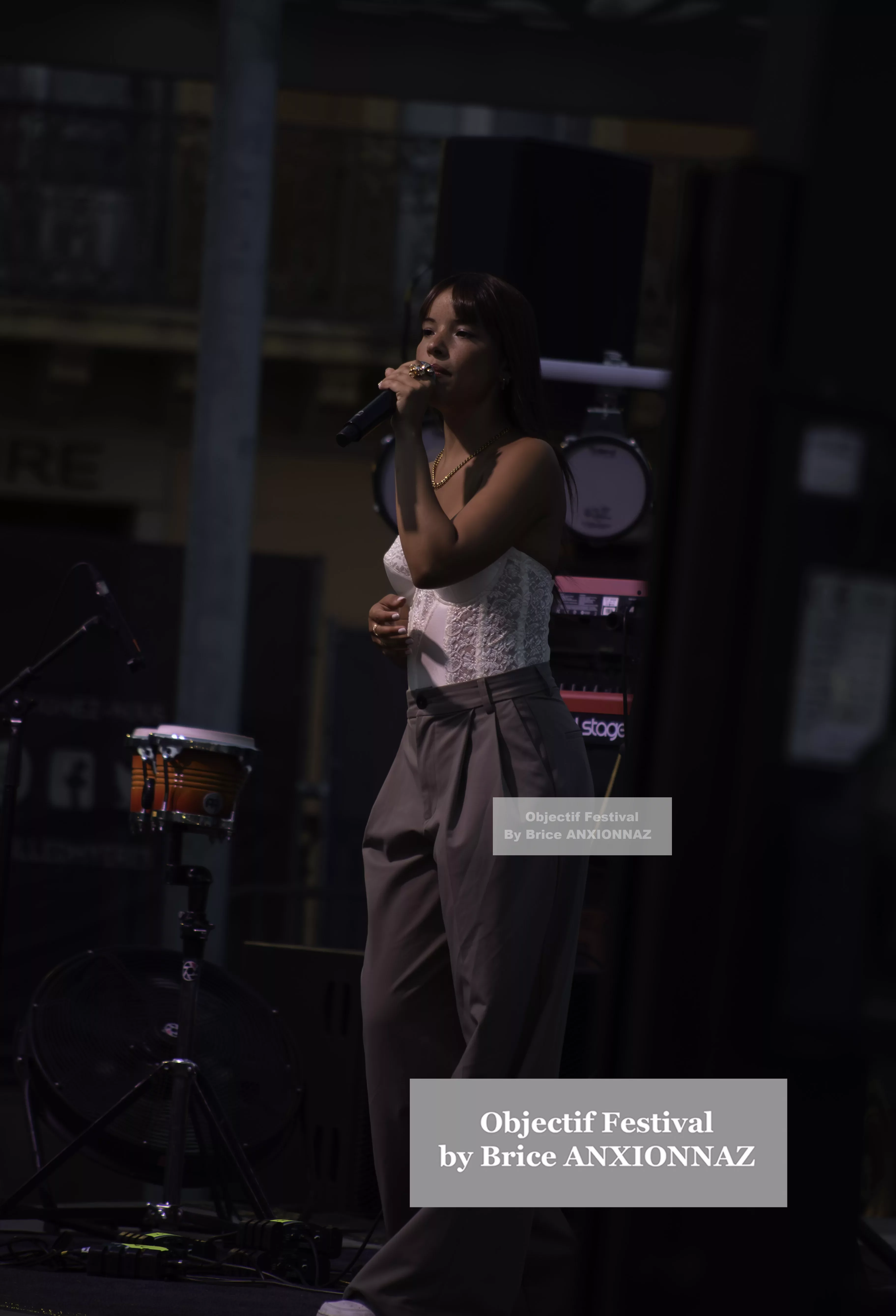 Une personne chante sur scène, vêtue d'un haut blanc et d'un pantalon gris, avec des instruments de musique en arrière-plan, dans une ambiance de festival. Itsouidad, Hyeres (06/08/2024)
