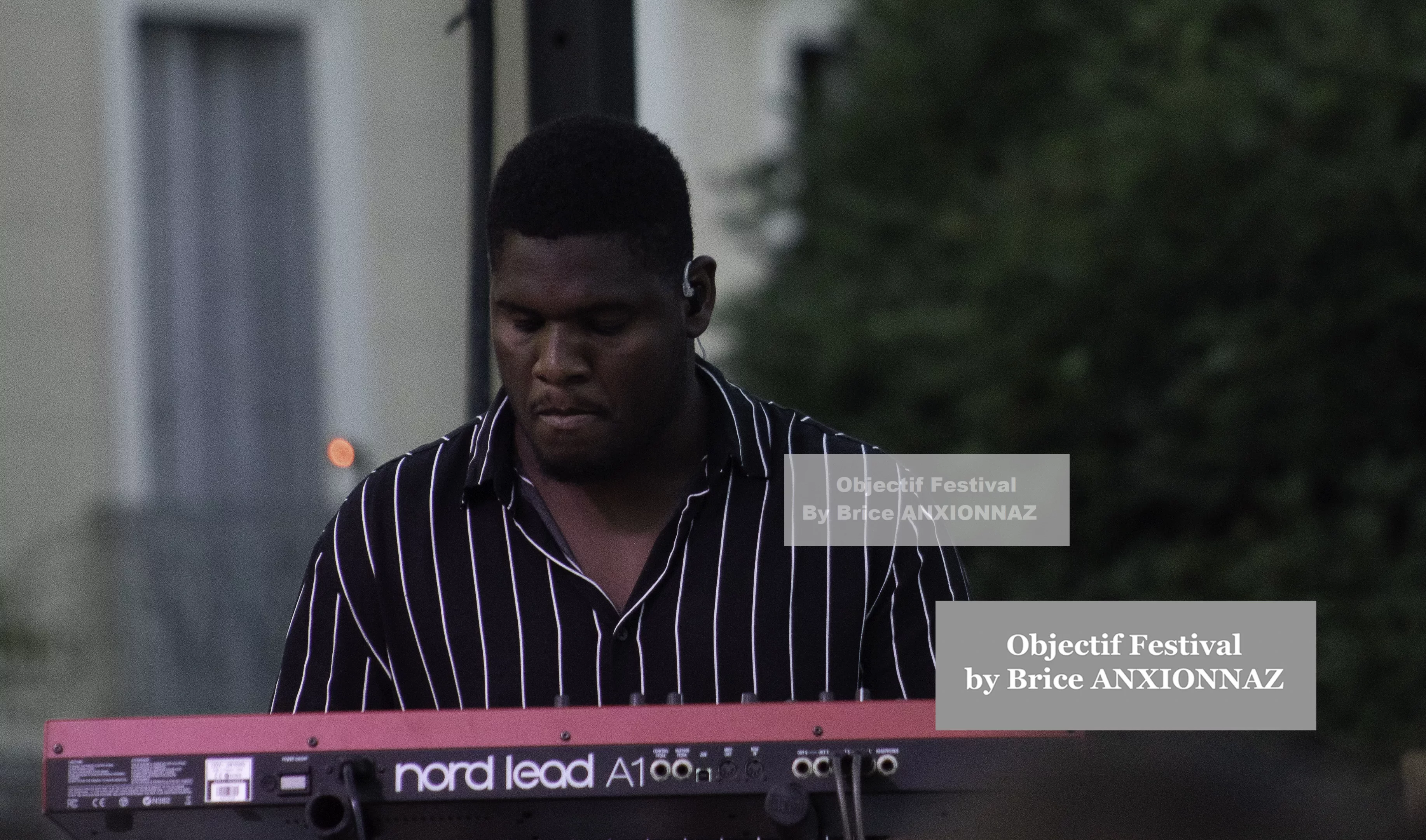 Un musicien joue du clavier sur scène, vêtu d'une chemise à rayures noires et blanches, avec un fond flou de verdure et une ambiance de festival en soirée. Itsouidad, Hyeres (06/08/2024)