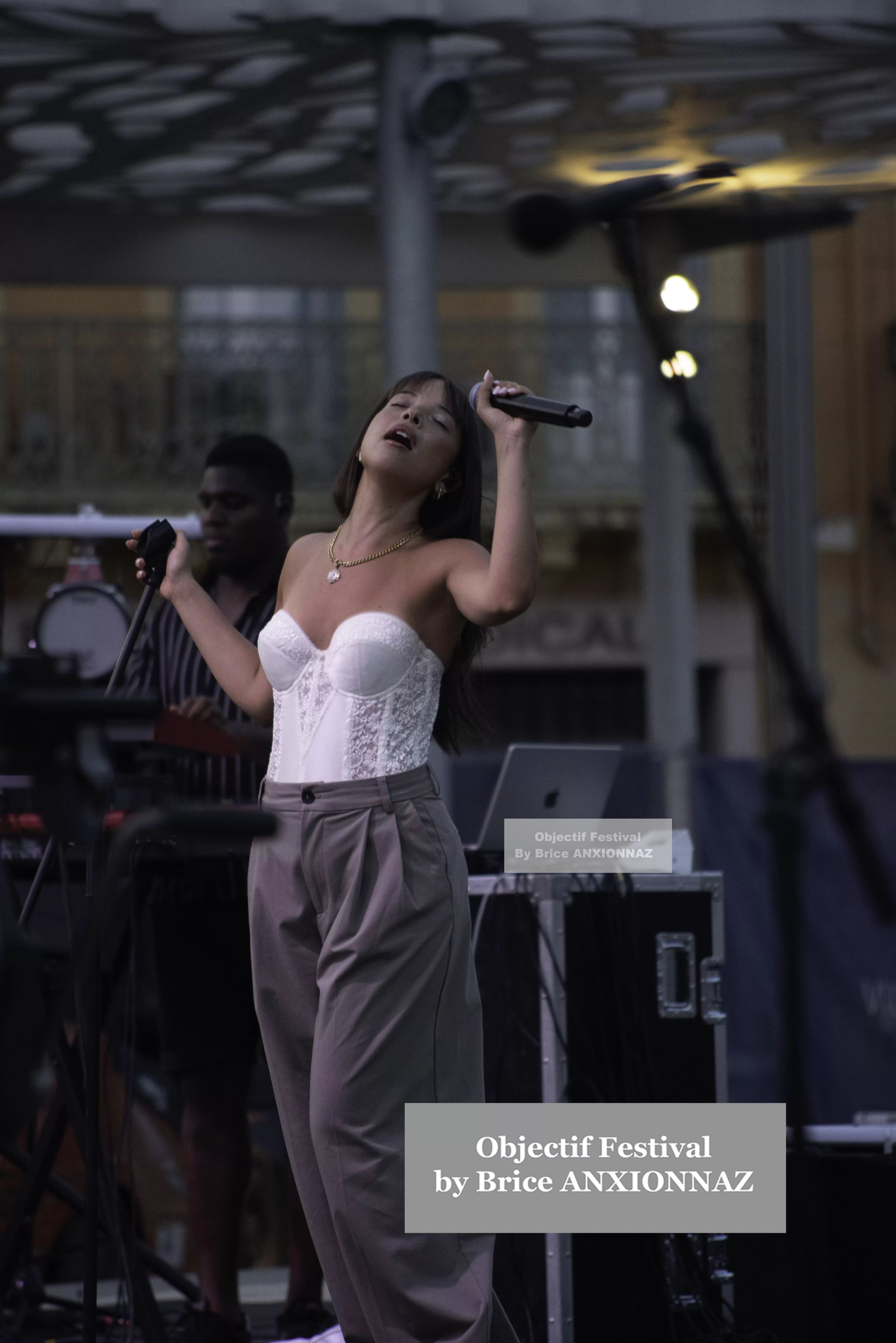 Une artiste chante sur scène, vêtue d'un haut blanc et de pantalons gris, tandis qu'un musicien l'accompagne en arrière-plan, sous une lumière douce et une ambiance festive. Itsouidad, Hyeres (06/08/2024)