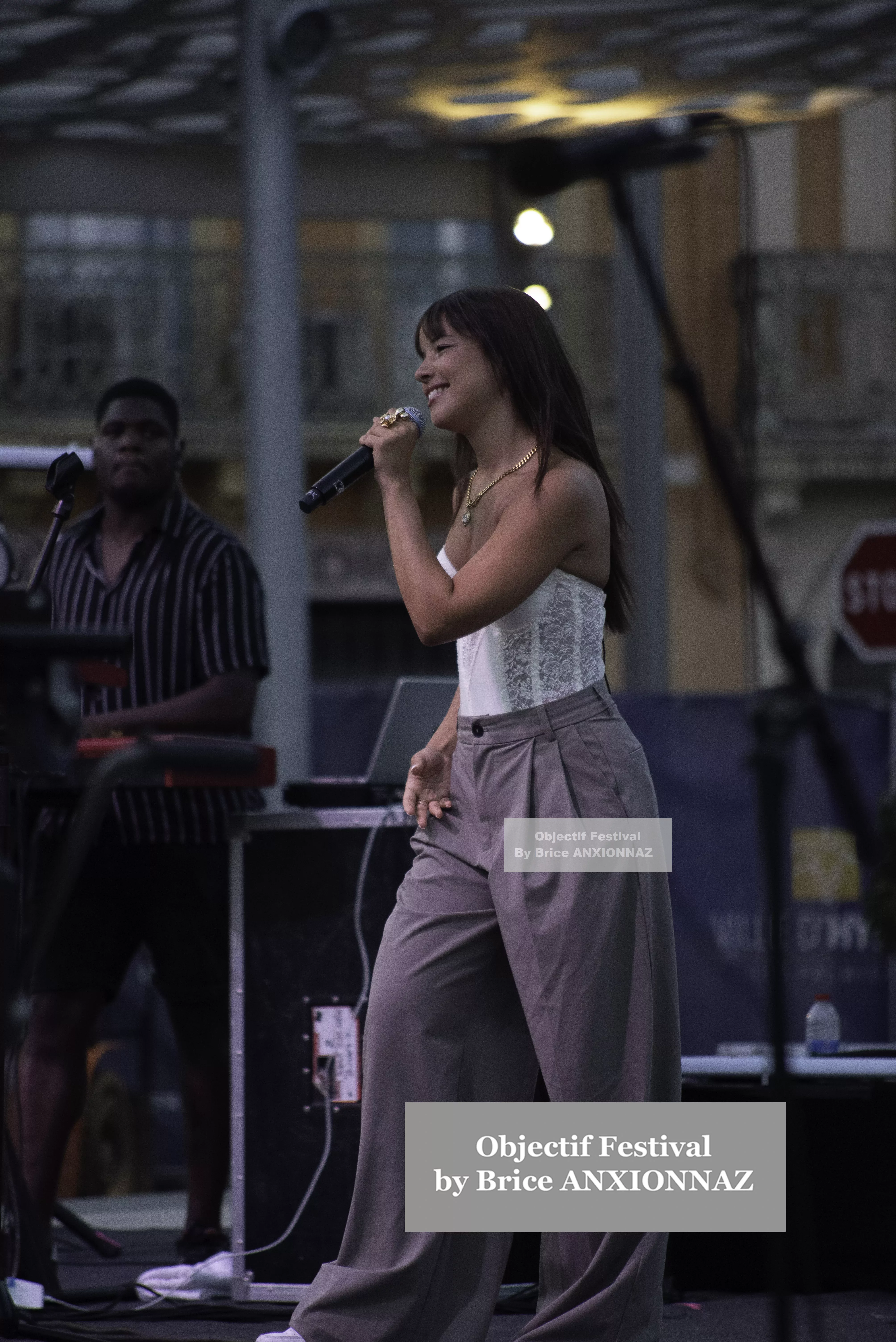 Une chanteuse se produit sur scène, vêtue d'un haut blanc et de pantalons gris, tandis qu'un musicien en arrière-plan joue d'un instrument, sous une lumière douce et un ciel dégagé. Itsouidad, Hyeres (06/08/2024)
