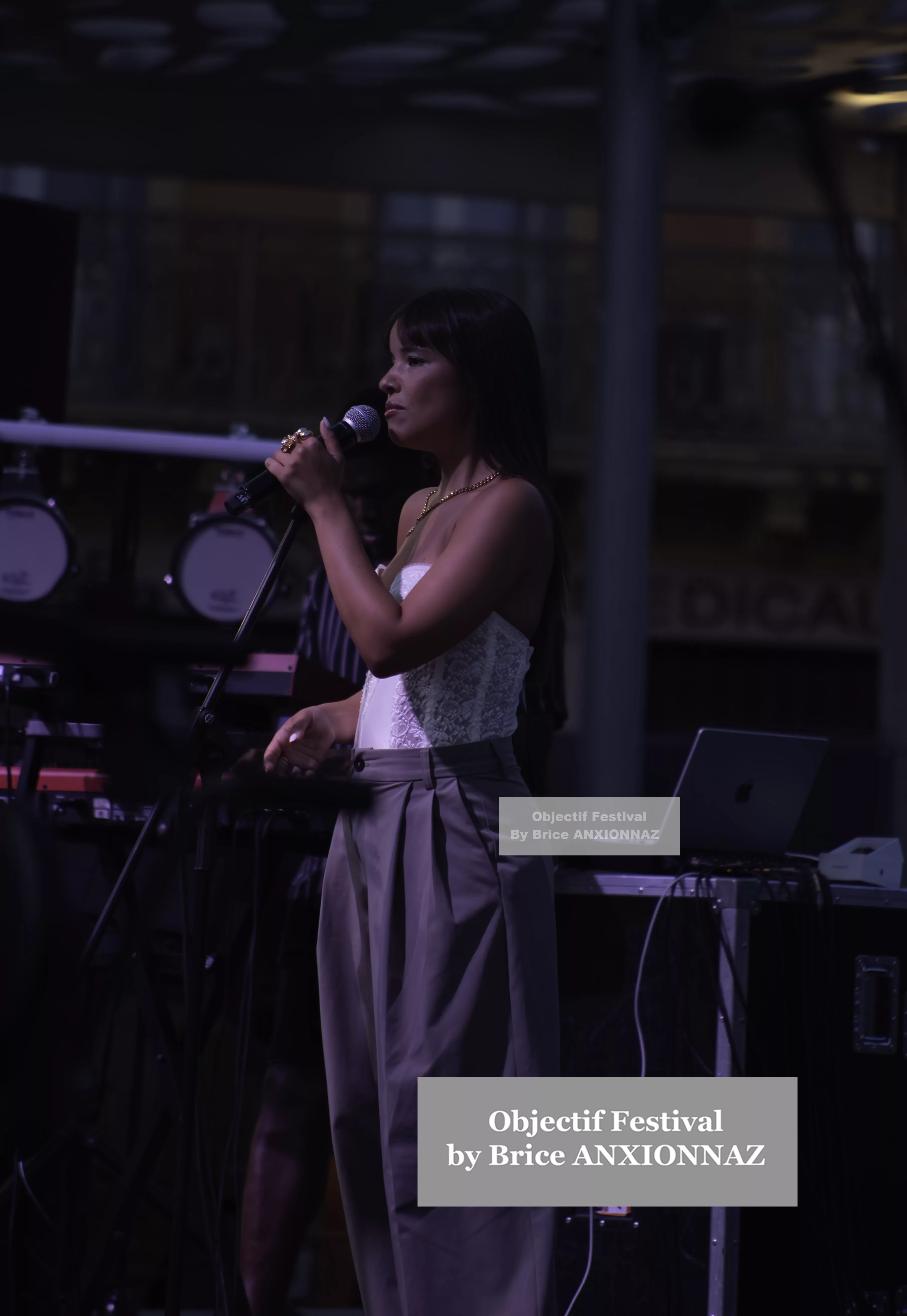 Une personne chante sur scène, vêtue d'un haut blanc et d'un pantalon gris, avec un microphone à la main, entourée d'équipements musicaux dans une ambiance tamisée. Itsouidad, Hyeres (06/08/2024)
