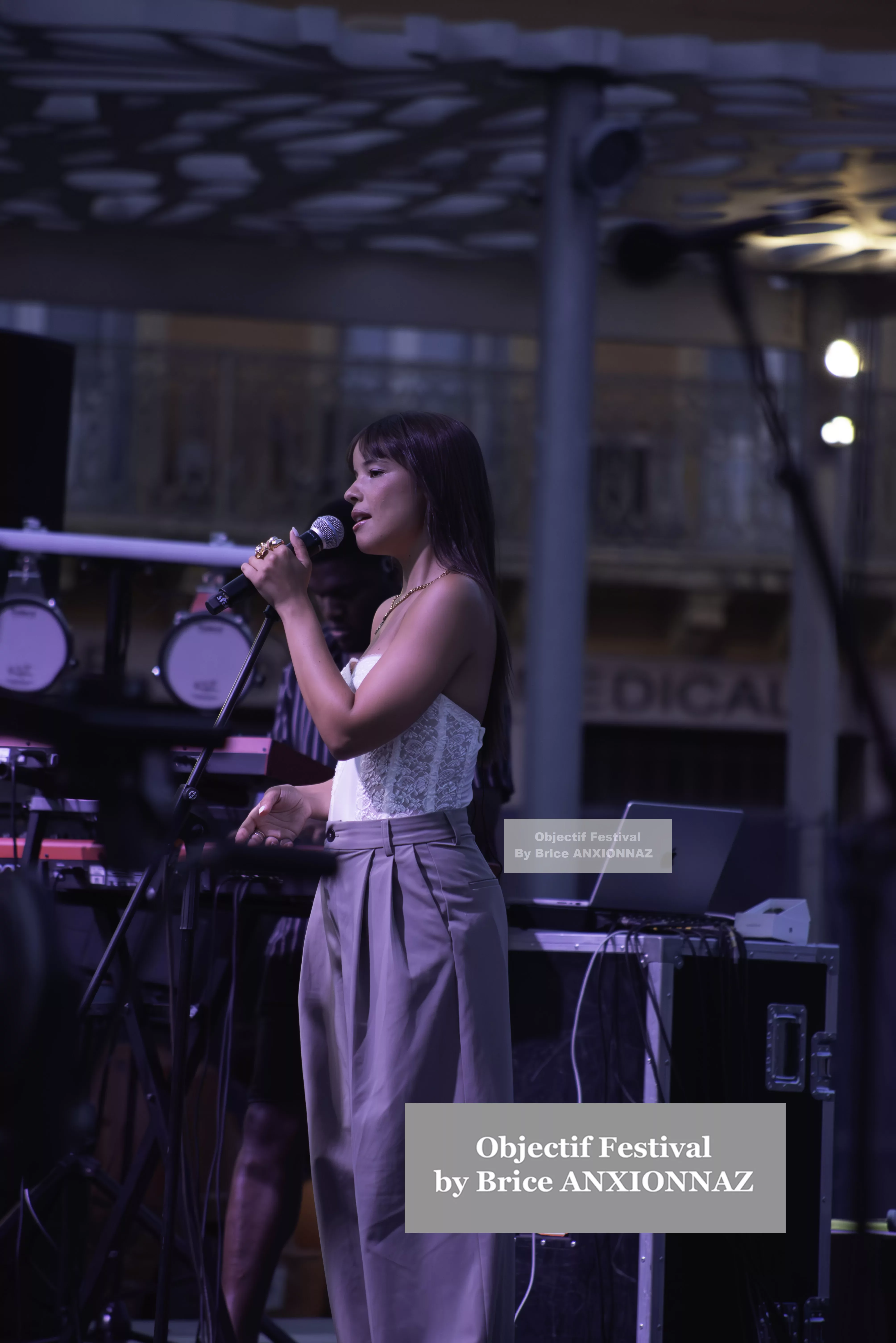 Une chanteuse se produit sur scène, vêtue d'un haut blanc et d'un pantalon gris, avec des instruments de musique en arrière-plan, sous un éclairage tamisé. Itsouidad, Hyeres (06/08/2024)