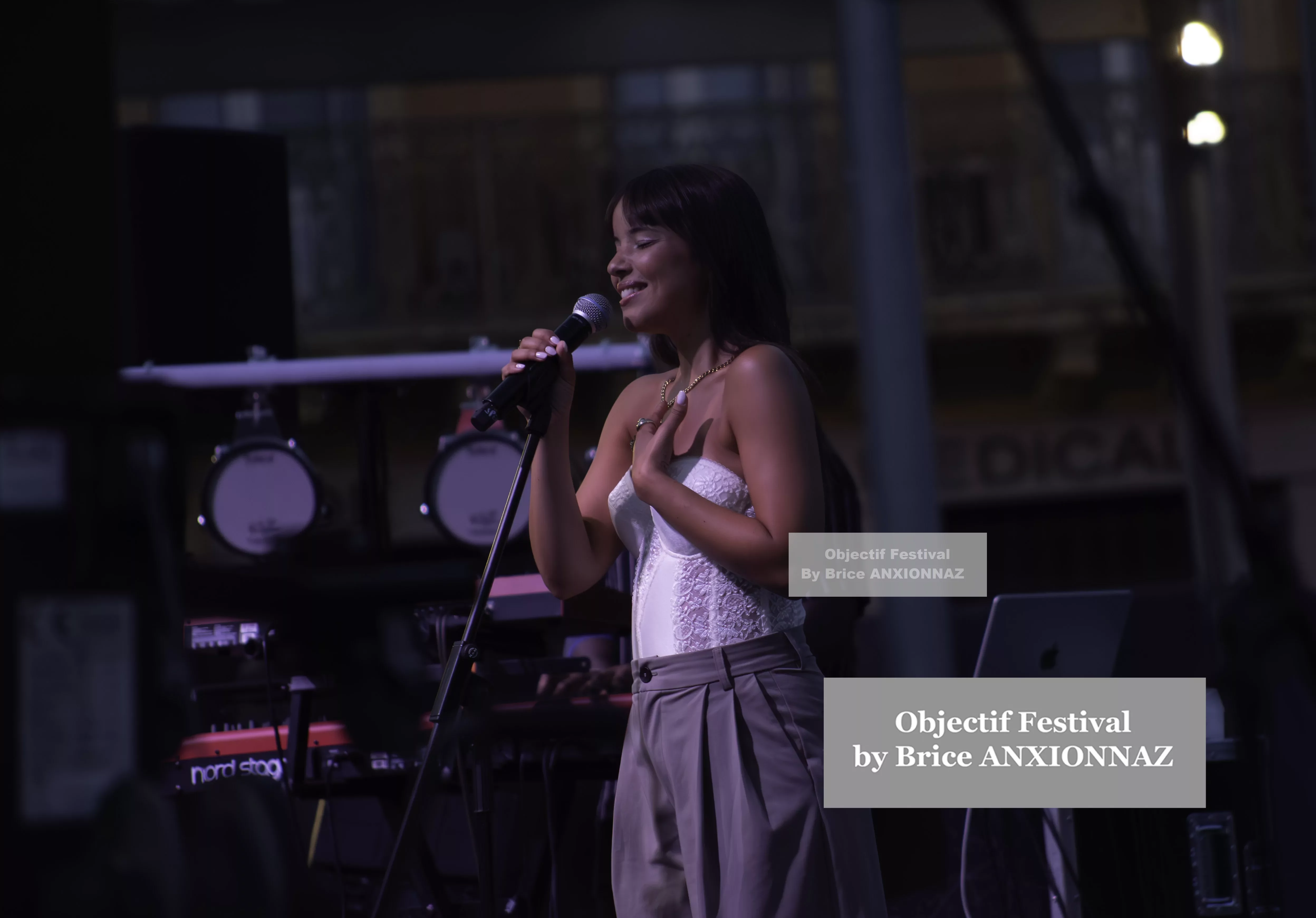 Une personne chante sur scène, vêtue d'un haut blanc et d'un pantalon gris, avec un microphone à la main, sous des lumières tamisées, créant une ambiance festive. Itsouidad, Hyeres (06/08/2024)
