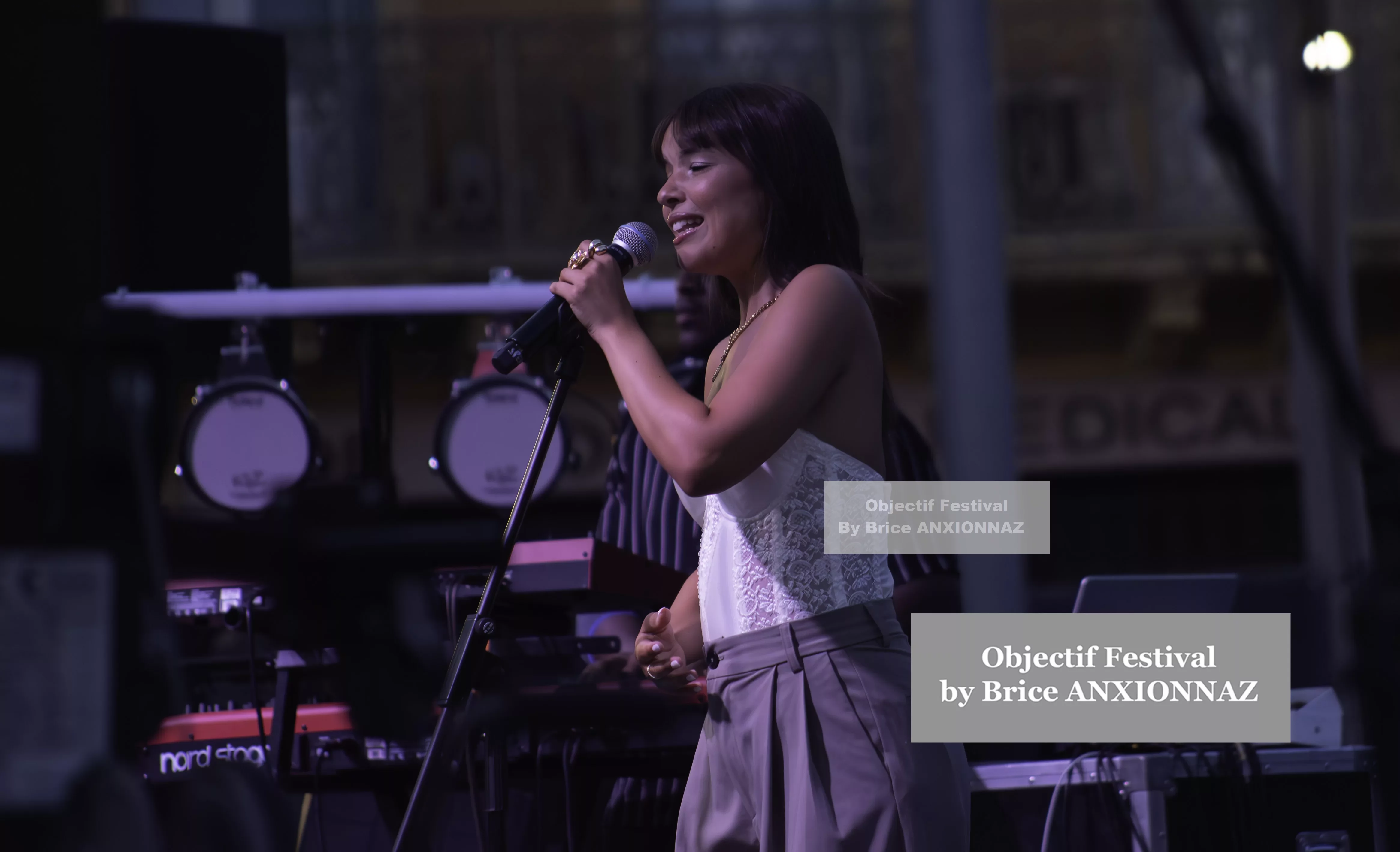 Une chanteuse se produit sur scène, vêtue d'un haut blanc et d'un pantalon clair, avec des instruments de musique en arrière-plan, sous une lumière tamisée. Itsouidad, Hyeres (06/08/2024)