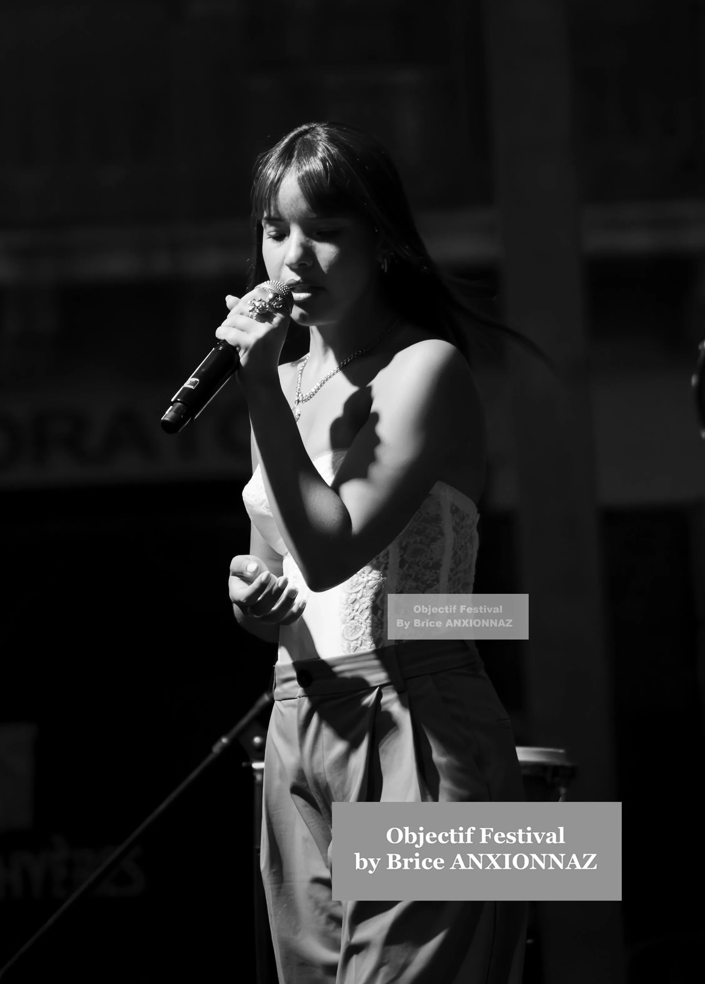 Une personne chante sur scène, vêtue d'un haut blanc et d'un pantalon clair, sous un éclairage contrasté en noir et blanc, créant une ambiance dynamique. Itsouidad, Hyeres (06/08/2024)