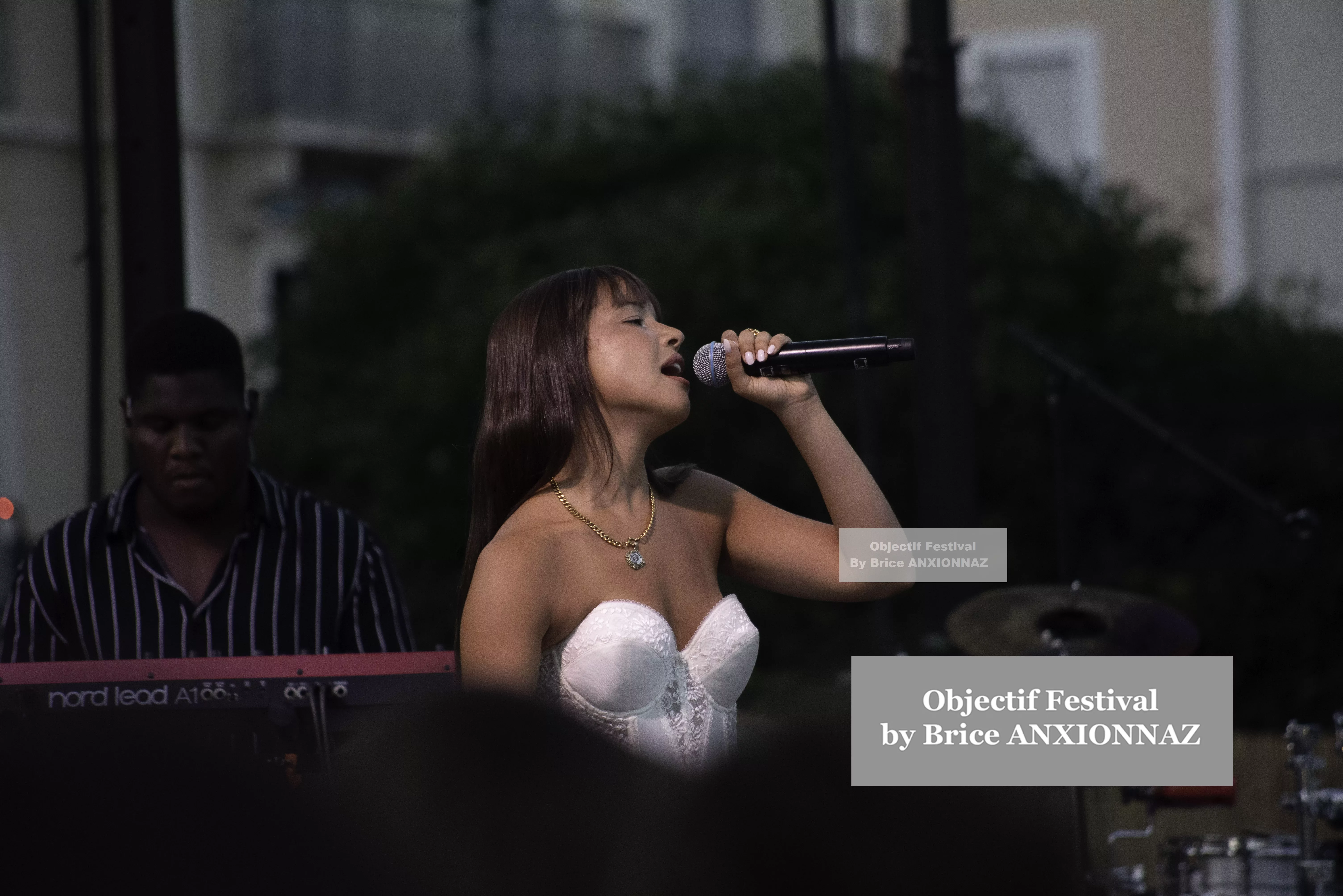 Une chanteuse en robe blanche se produit sur scène, microphone à la main, tandis qu'un musicien joue du clavier en arrière-plan, sous une lumière douce et une ambiance festive. Itsouidad, Hyeres (06/08/2024)