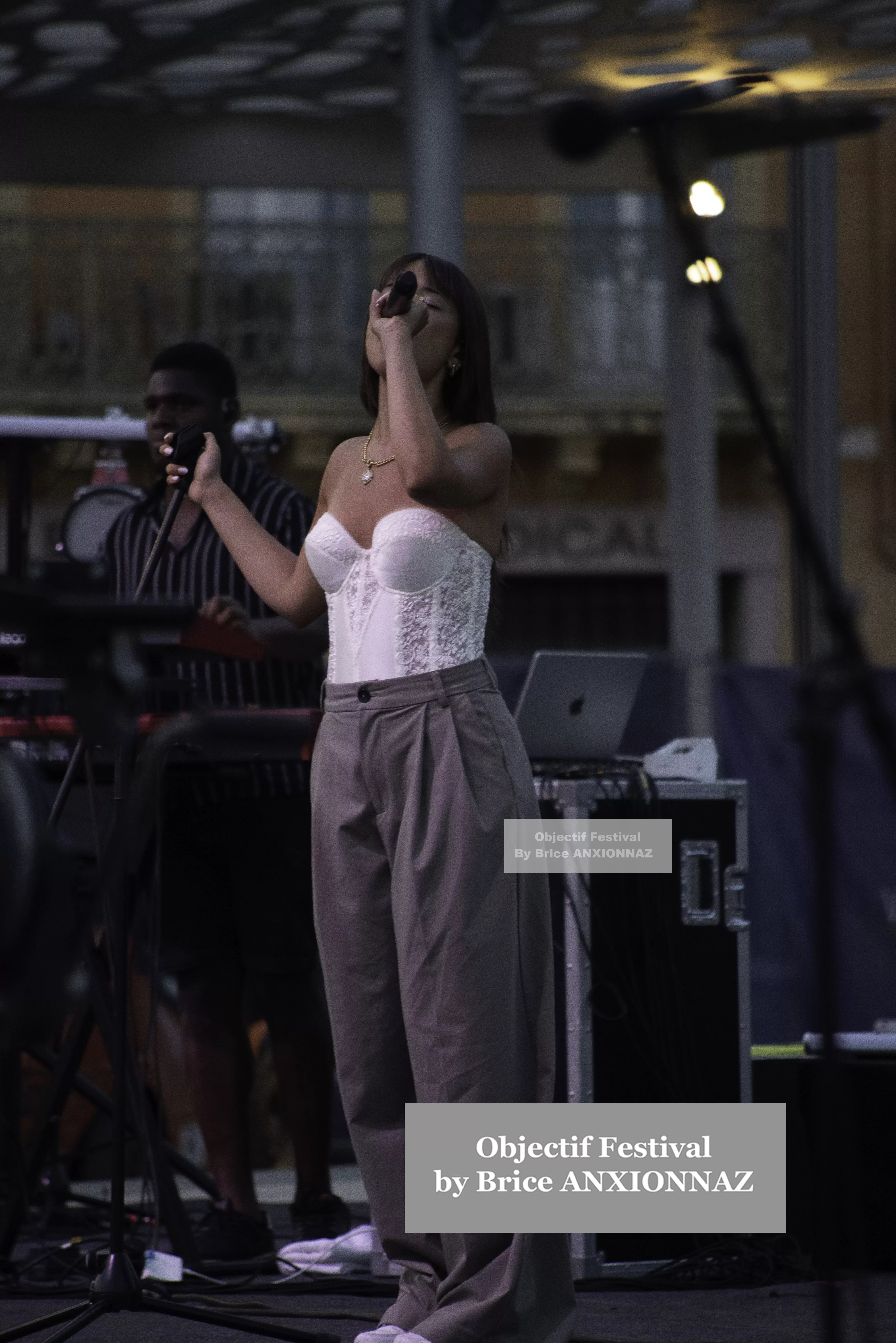 Une artiste chante sur scène, vêtue d'un haut blanc et de pantalons larges, tandis qu'un musicien l'accompagne en arrière-plan, créant une ambiance festive sous un éclairage doux. Itsouidad, Hyeres (06/08/2024)