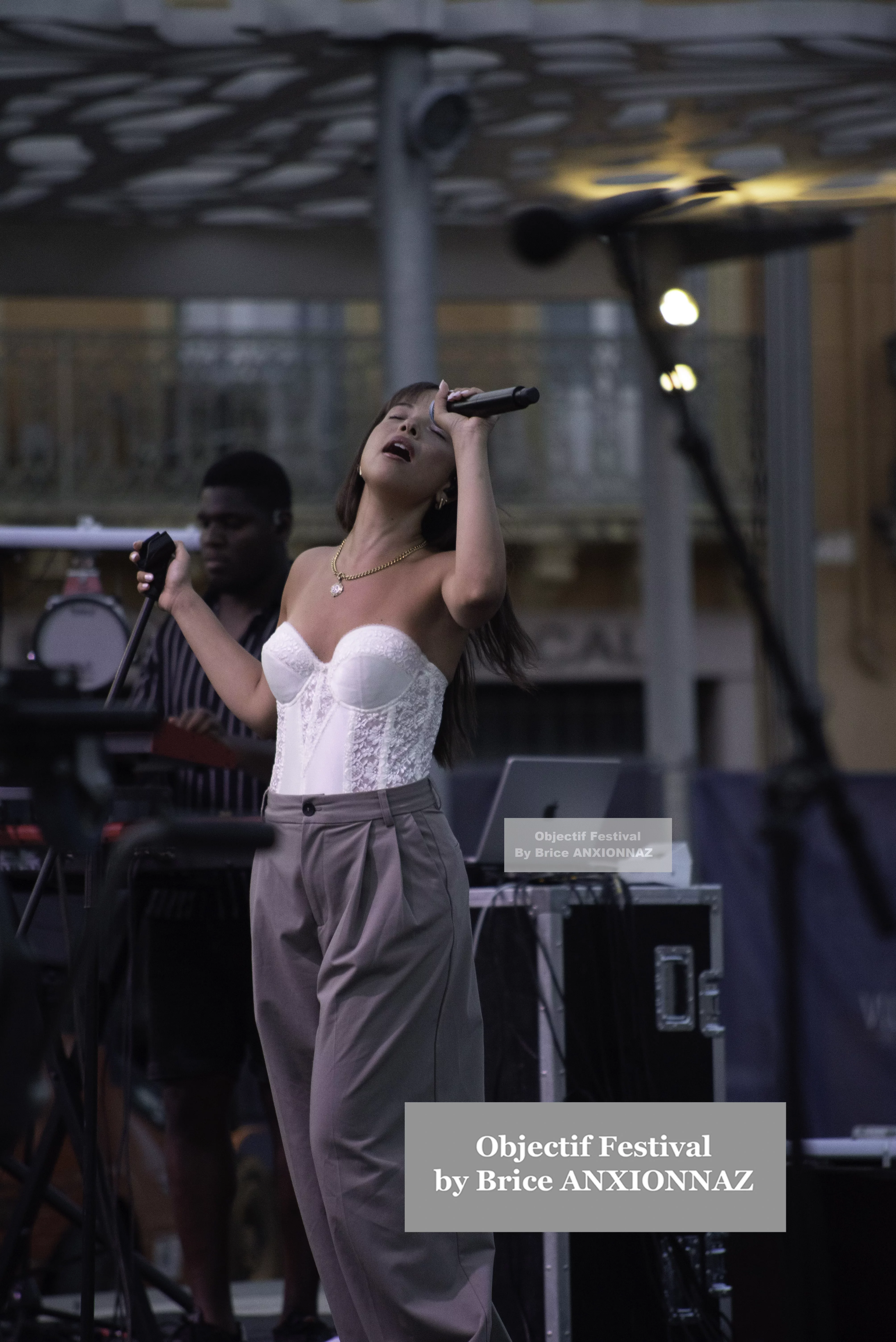 Une artiste chante sur scène, vêtue d'un haut blanc et de pantalons gris, tandis qu'un musicien en arrière-plan joue d'un instrument, sous une lumière douce et une ambiance festive. Itsouidad, Hyeres (06/08/2024)
