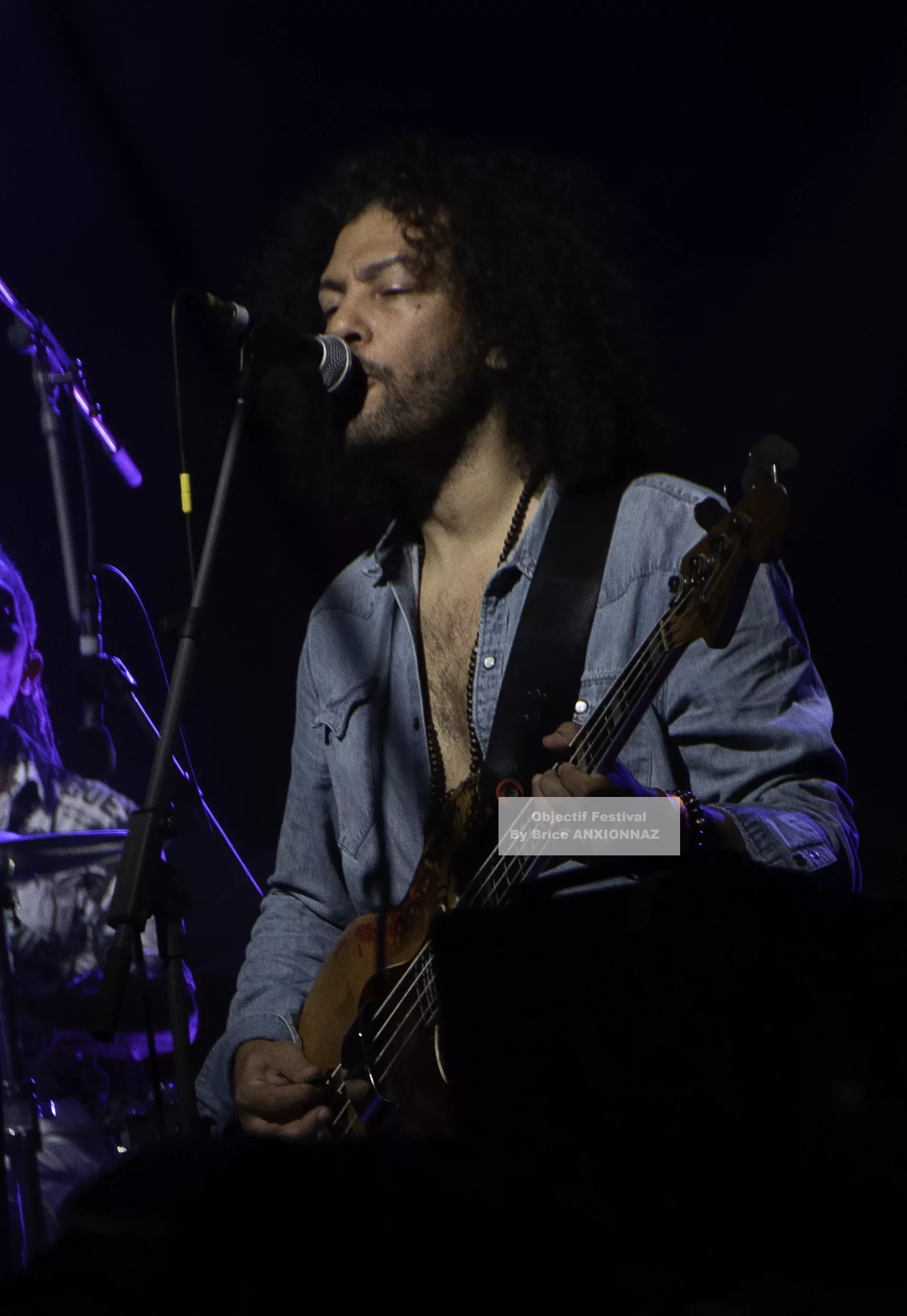 Louis Bertignac — Un musicien joue de la basse sur scène, vêtu d'une chemise en denim, avec des lumières tamisées et une ambiance dynamique, tandis qu'un batteur est visible en arrière-plan. 21 07 2026, Hyeres (21/07/2026)
