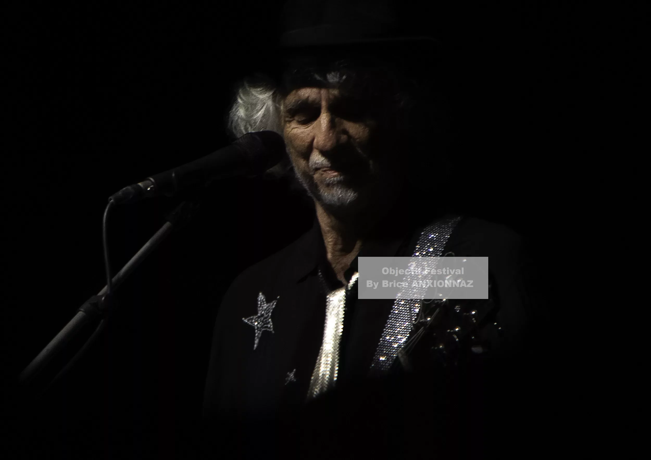 Louis Bertignac — Louis Bertignac — Un individu chante sur scène, vêtu d'une chemise noire ornée d'étoiles et d'une bandoulière scintillante, dans une ambiance sombre et intimiste, avec un éclairage focalisé.