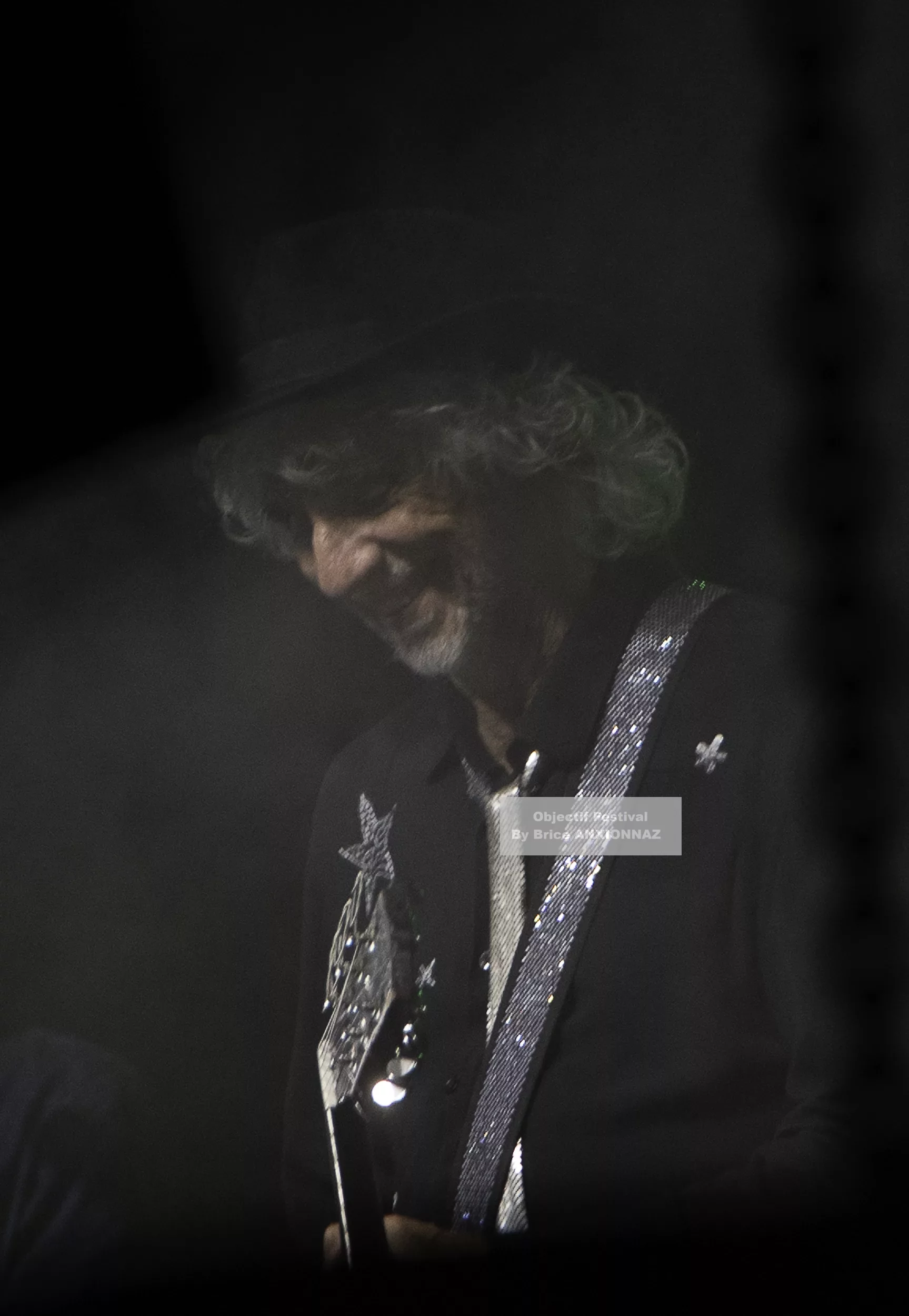 Louis Bertignac — Louis Bertignac — Un musicien souriant, vêtu d'une chemise noire ornée de paillettes, joue de la guitare dans une ambiance brumeuse, avec des lumières tamisées créant une atmosphère intimiste.