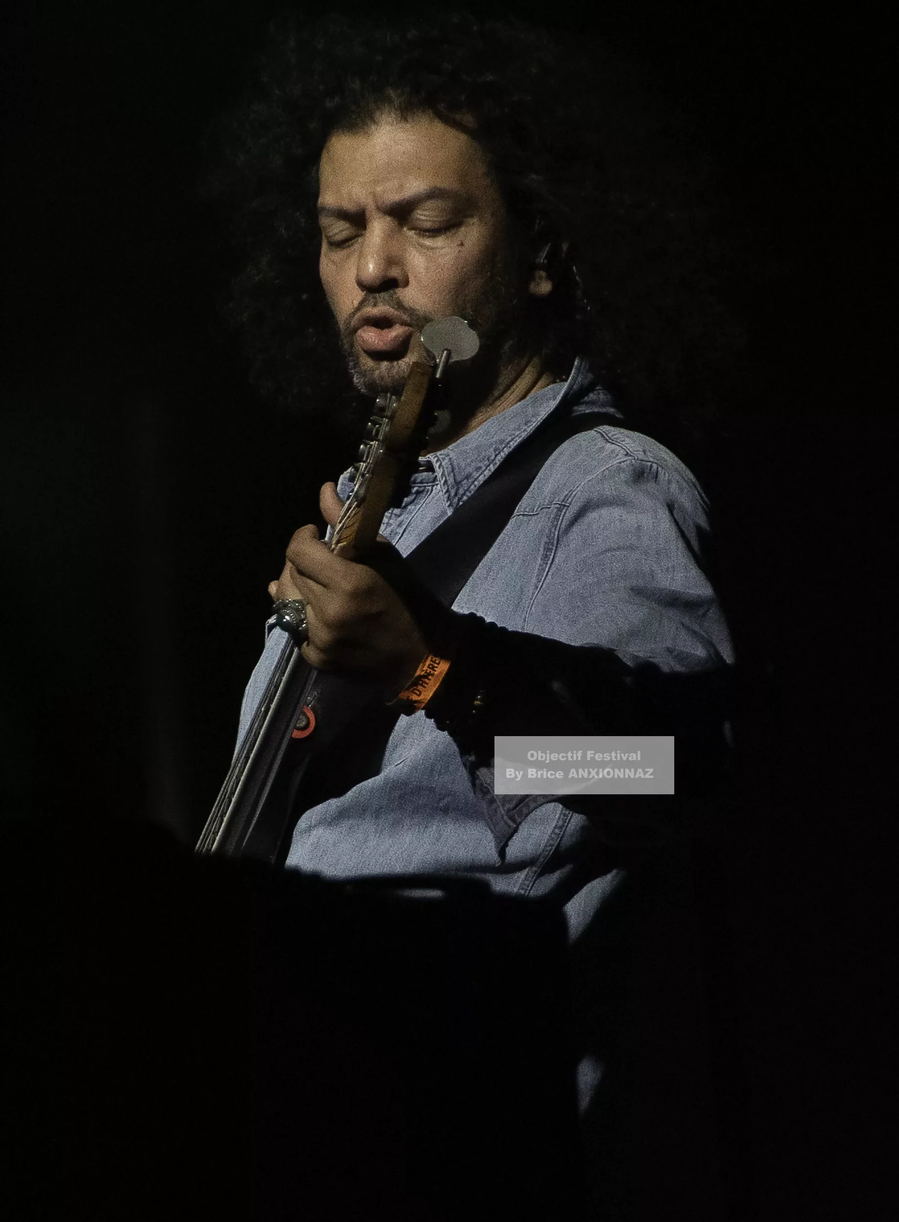 Louis Bertignac — Un musicien joue de la basse sur scène, éclairé par une lumière tamisée, portant une chemise en denim, avec des cheveux bouclés et une expression concentrée. 21 07 2026, Hyeres (21/07/2026)
