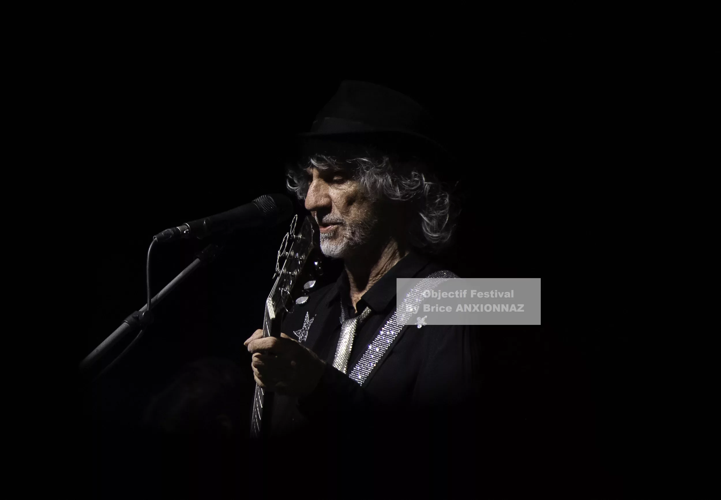 Louis Bertignac — Un homme avec des cheveux bouclés et un chapeau noir joue de la guitare sur scène, éclairé par une lumière douce sur fond sombre, créant une ambiance intimiste. 21 07 2026, Hyeres (21/07/2026)