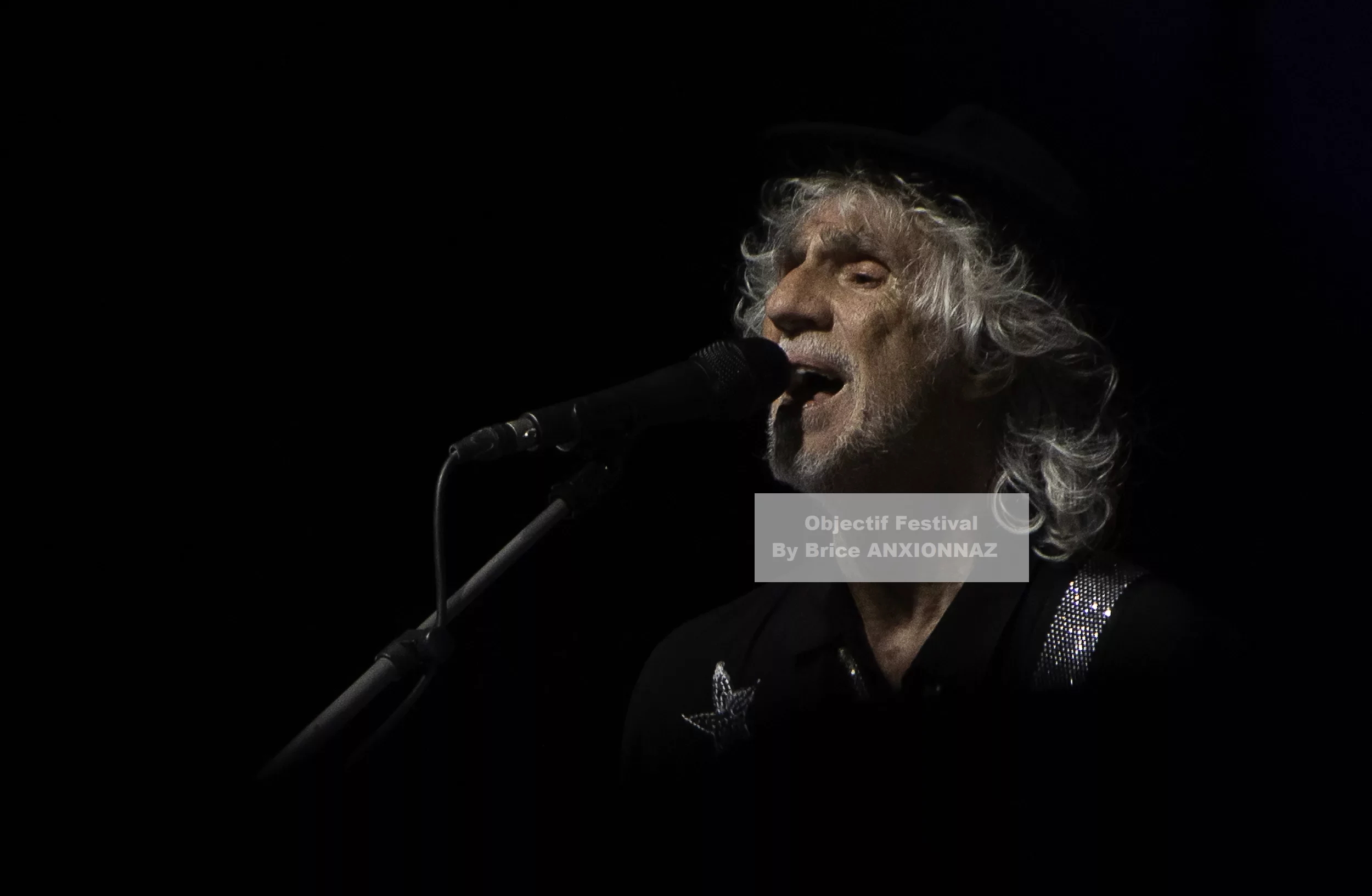 Louis Bertignac — Louis Bertignac — Un homme chante au microphone, éclairé par une lumière douce, avec des cheveux argentés et un chapeau noir, créant une ambiance intimiste sur fond sombre.